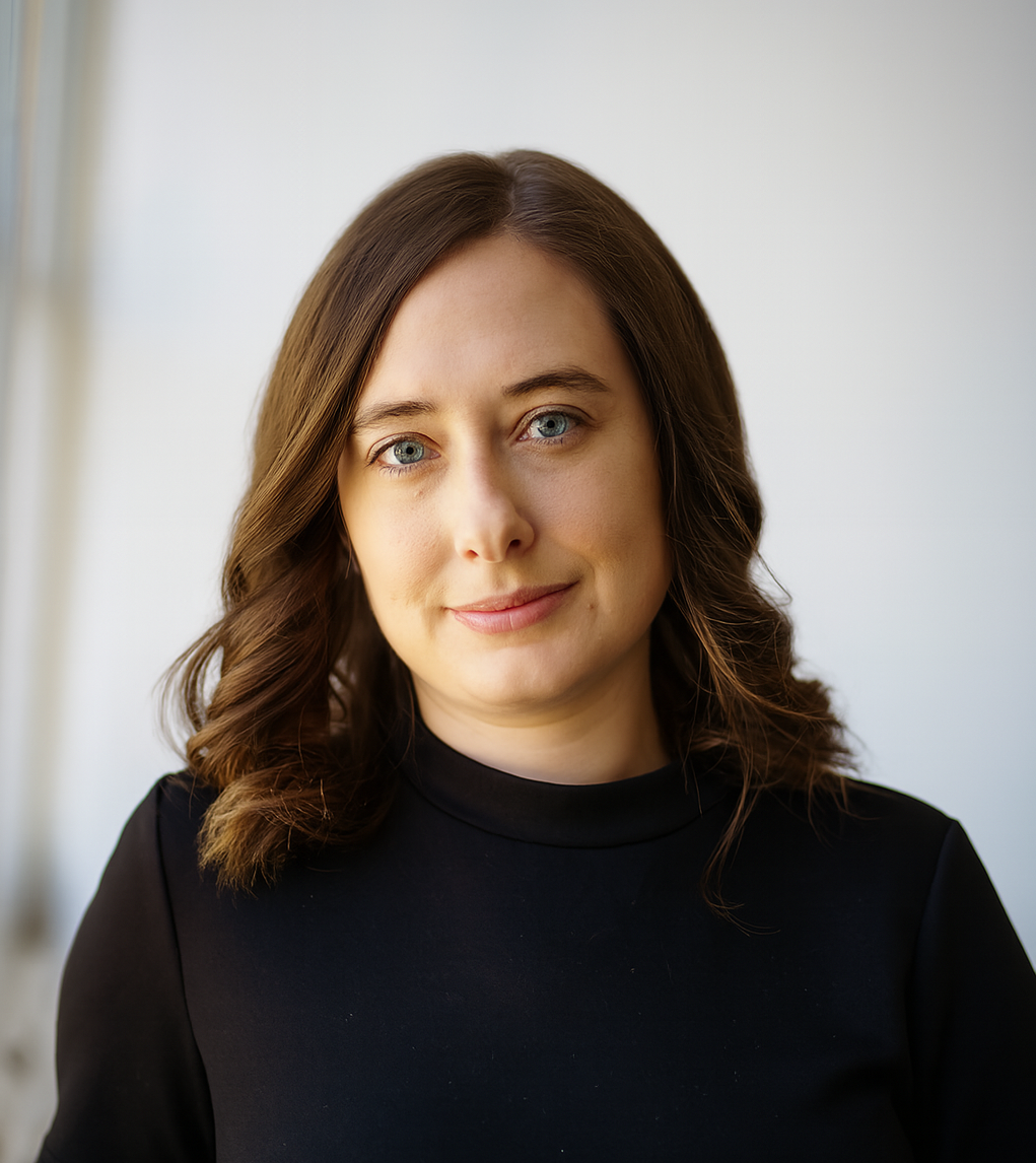 Headshot of a woman with brown hair and blue eyes, wearing a black top, in front of a plain light background.