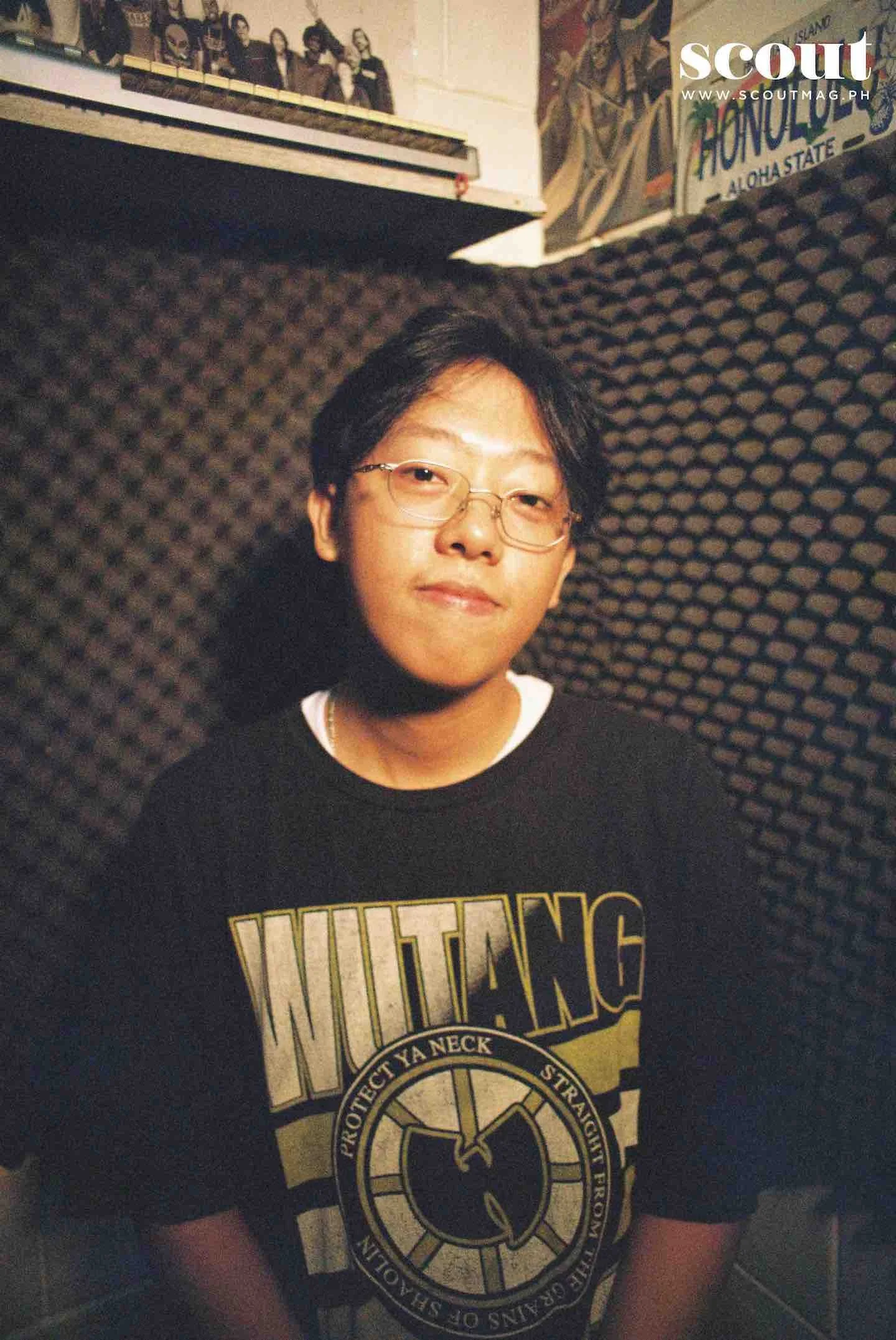 Waiian wearing a black T-shirt with yellow and white text that reads 'WU-TANG,' posing indoors with a textured soundproof wall behind him. There are posters and artwork visible in the background.