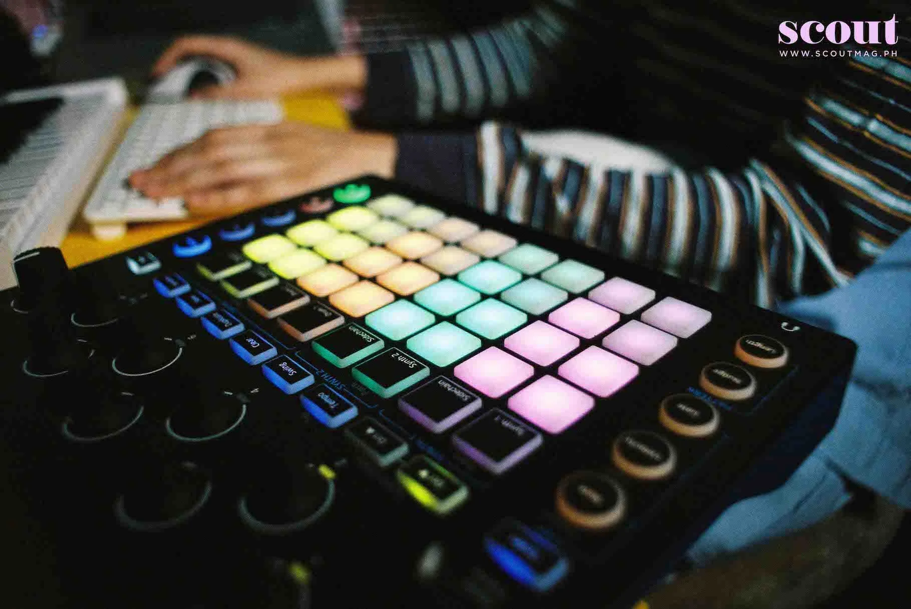 Pikoy working on a MIDI controller with LED pads glowing in various colors, with a keyboard in the background.