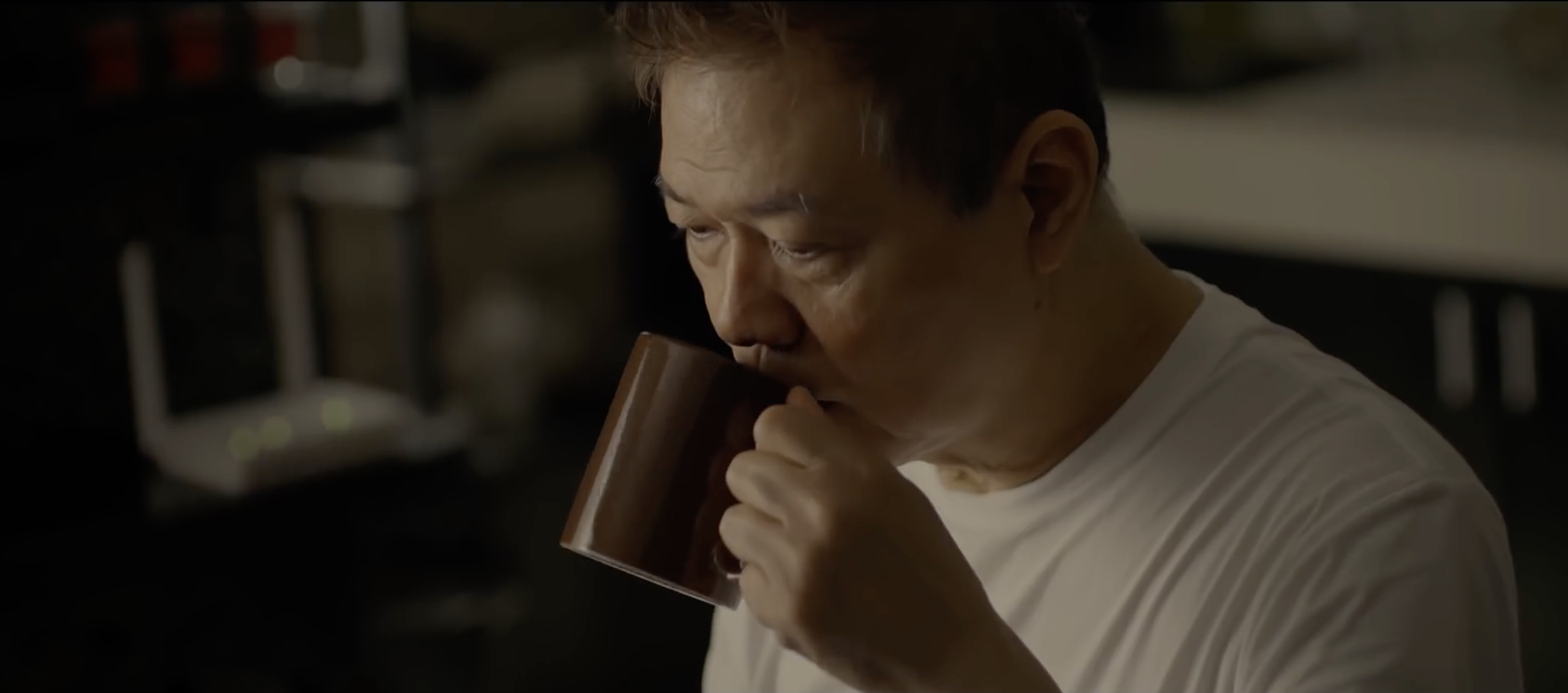 An older man with short hair and a white shirt drinks from a brown mug in a dimly lit room.