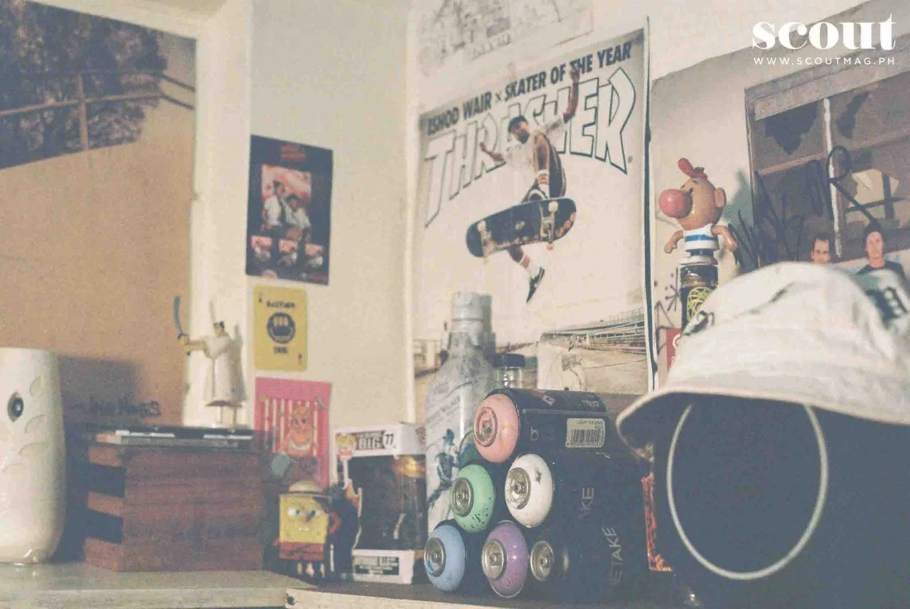 A desk or table with spray paint cans, artwork, and small figurines, including a character from comics, in a room decorated with posters and stickers.