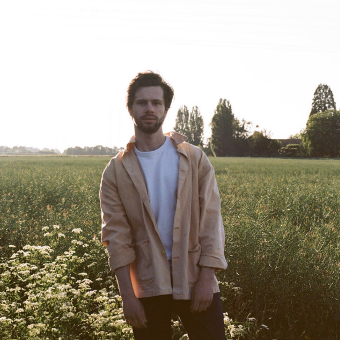 A young man with dark hair and a beard standing in a field during sunset, wearing a beige jacket over a white shirt.
