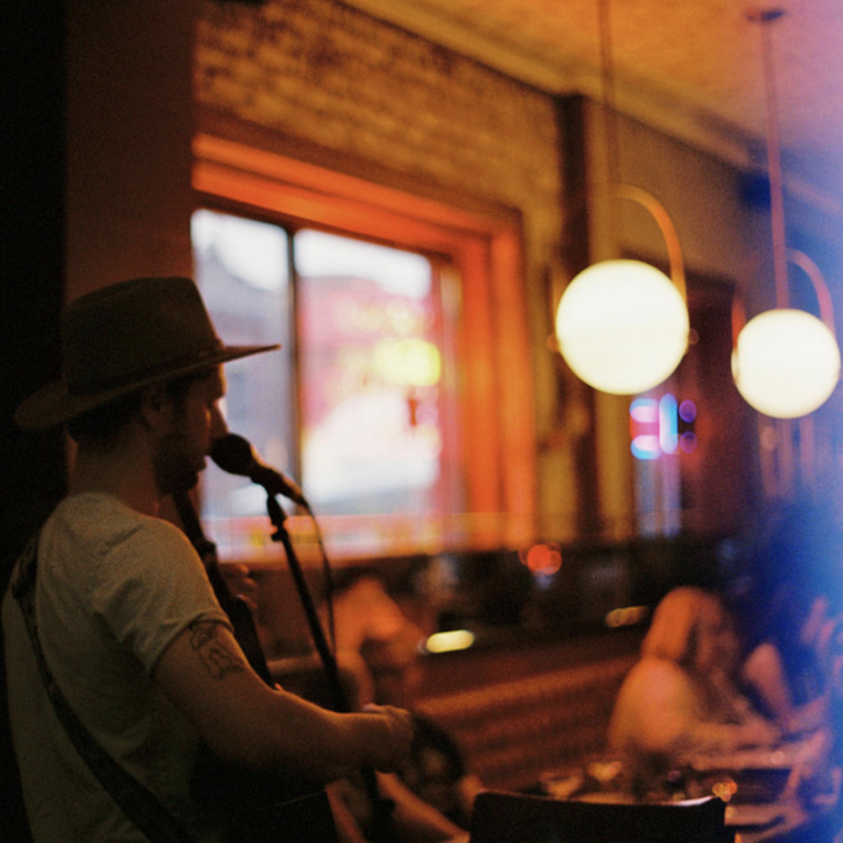 A musician wearing a hat sings into a microphone in a dimly lit, cozy bar with warm lights and neon signs visible outside the window.