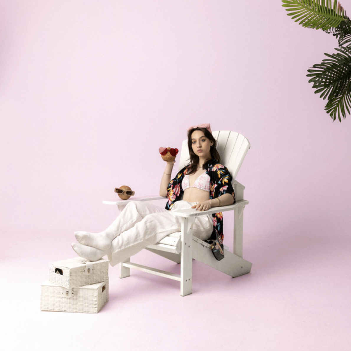 A woman sitting in a white lounge chair wearing a bikini top, a floral cover-up, and white pants, holding a pineapple-shaped drink with a cherry on top. She has a pink headband and is surrounded by summer accessories, with a pink background and green palm leaves in the corner.