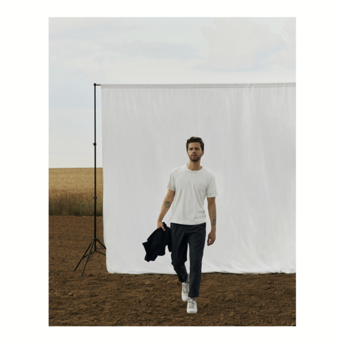 A man walking in front of a white backdrop outdoors, holding a black jacket, with a field and a cloudy sky in the background.