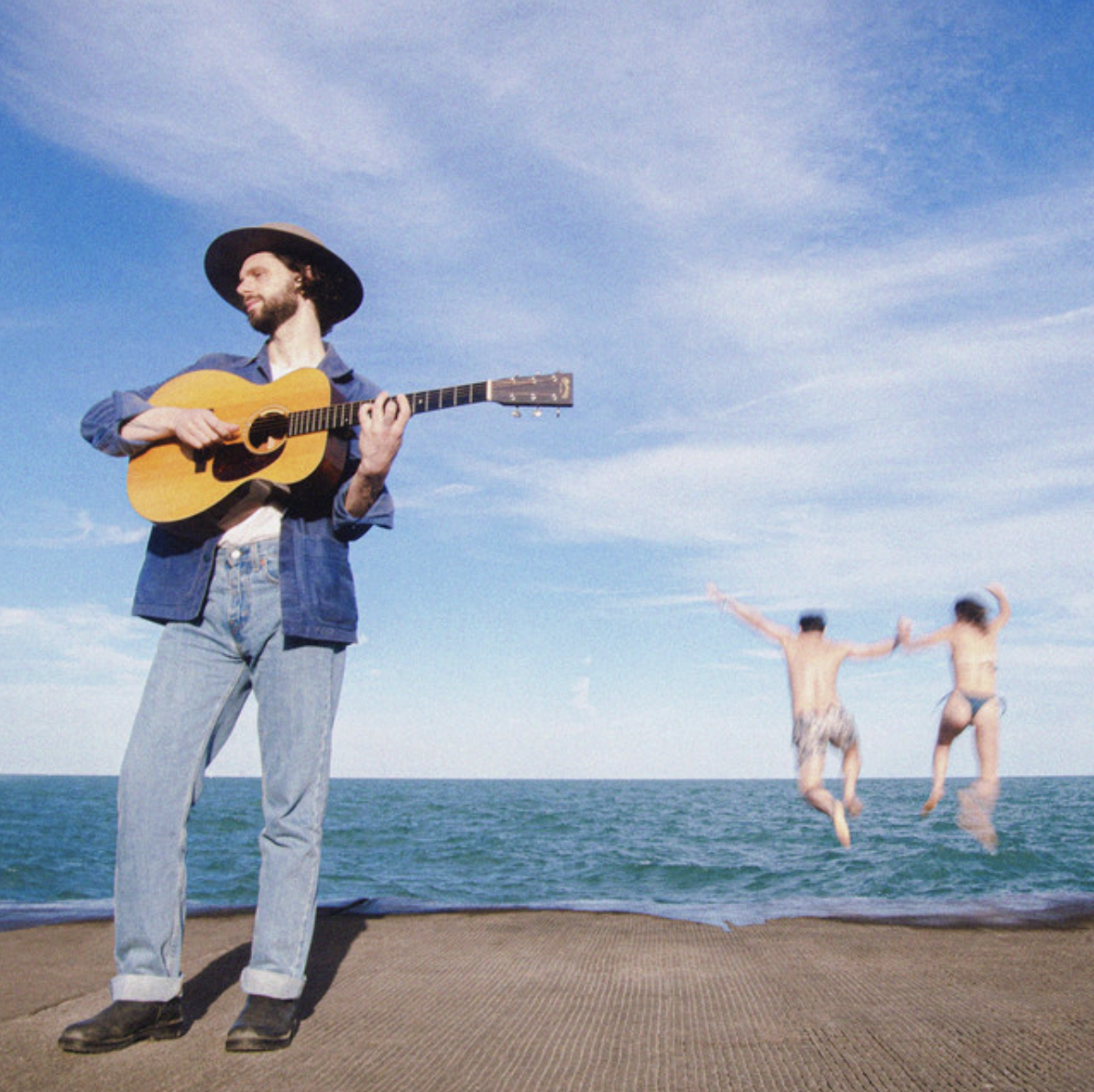 A man in a hat playing an acoustic guitar on a surface near the ocean, with two children jumping into the water in the background under a blue sky.