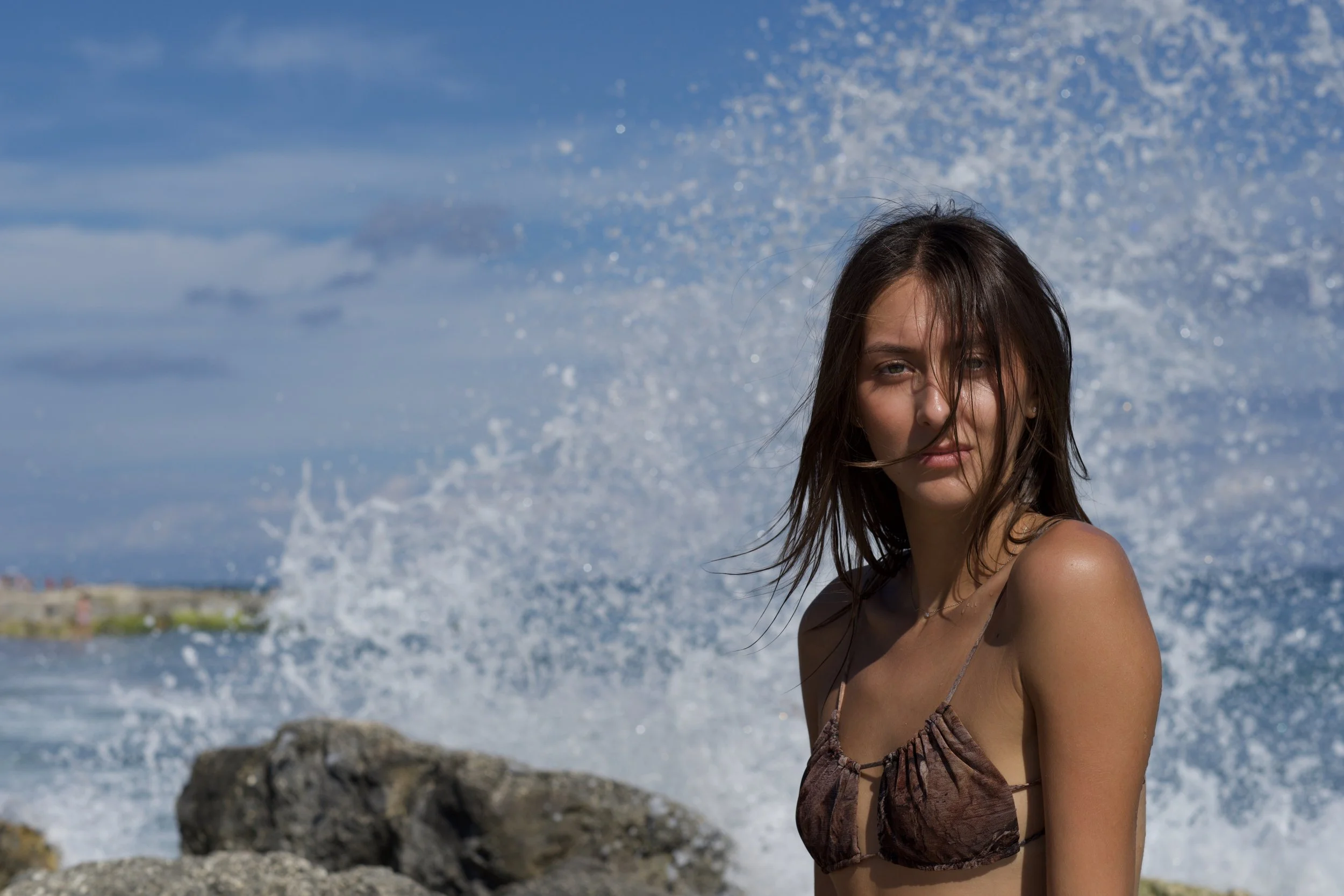 A bust-length portrait of Katia Mueller standing in front of the ocean in a brown bikini top as a wave crashes behind her, looking directly at the camera.