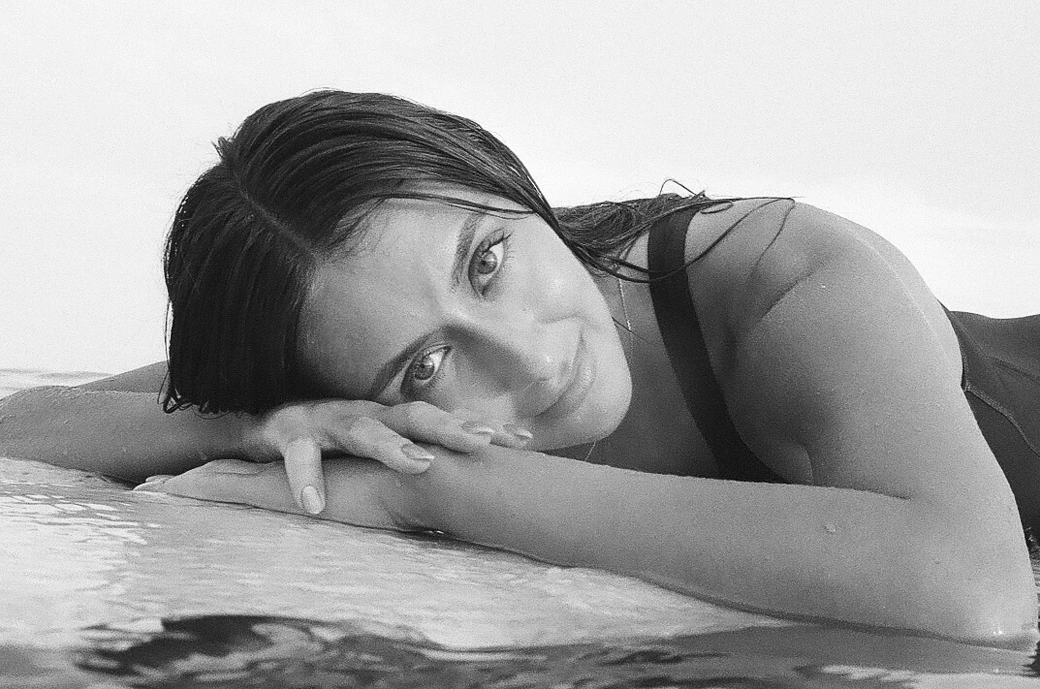 A close-up black-and-white headshot of Katia Mueller lying on the edge of a pool, looking directly at the camera.