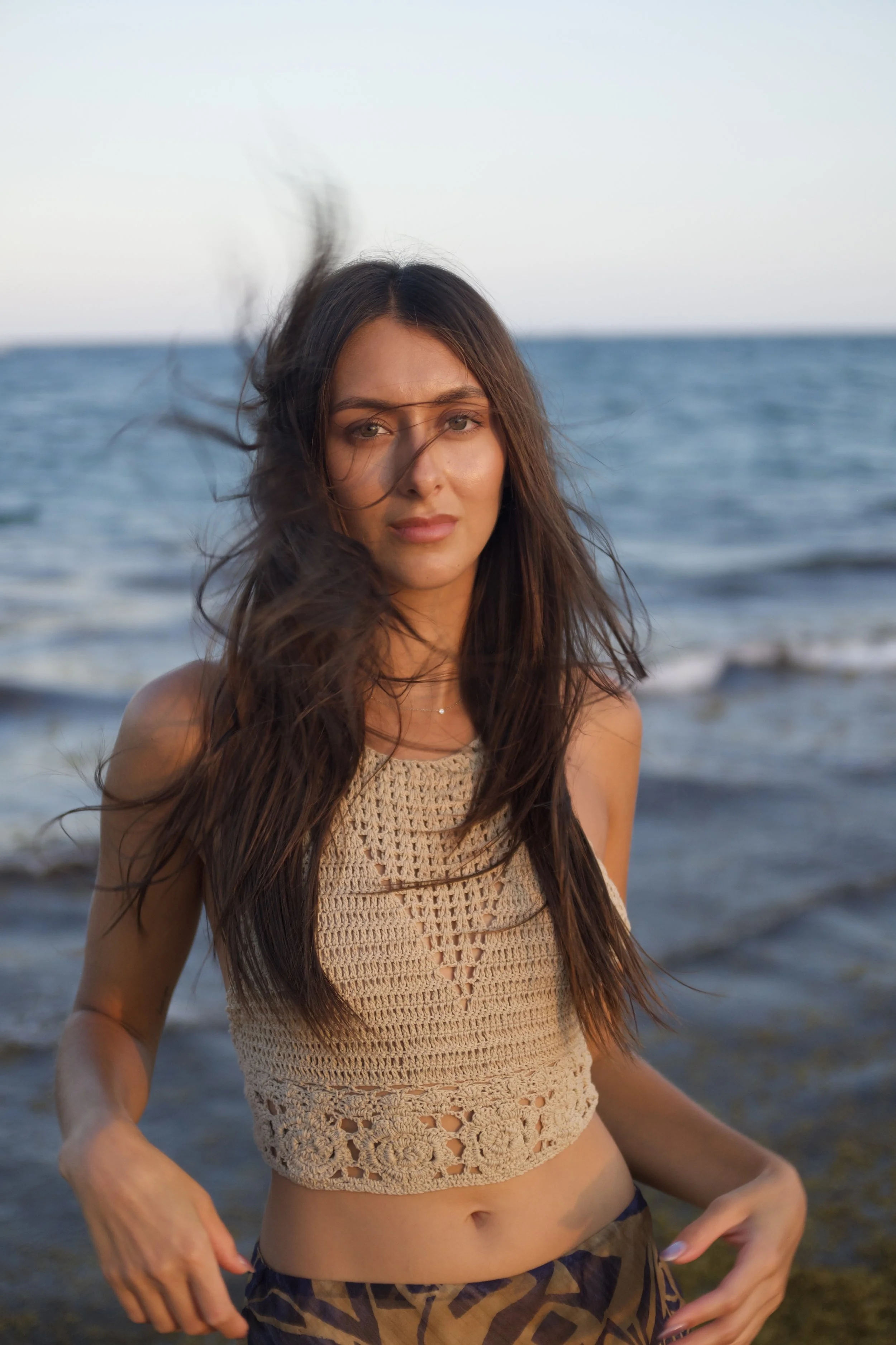A mid-length portrait of Katia Mueller standing in front of the ocean in a white bohemian top on a windy day, looking directly at the camera.