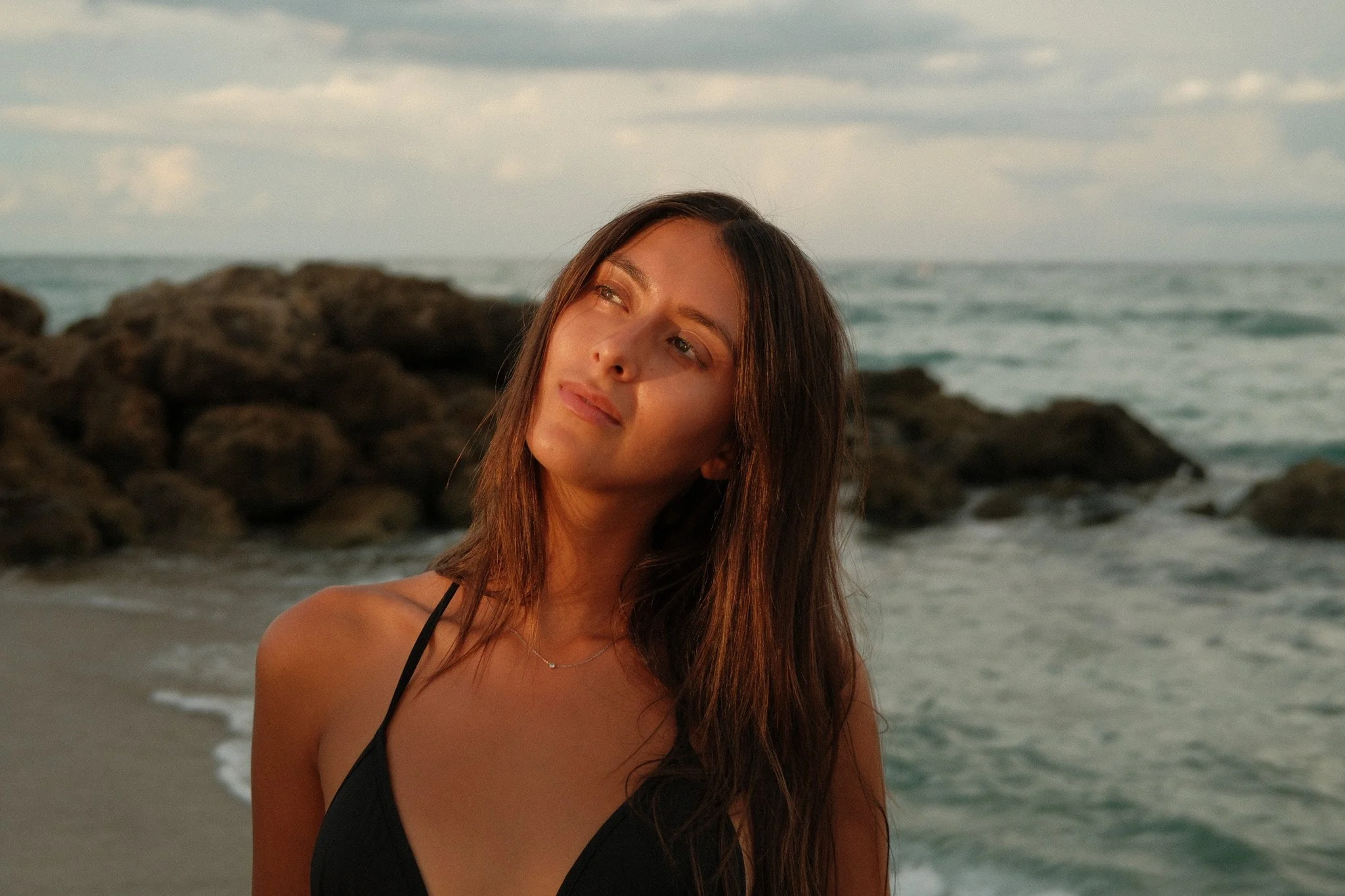 A close-up headshot of Katia Mueller standing on the beach at sunset in front of a rock formation on the shore, looking away from the camera at the horizon.