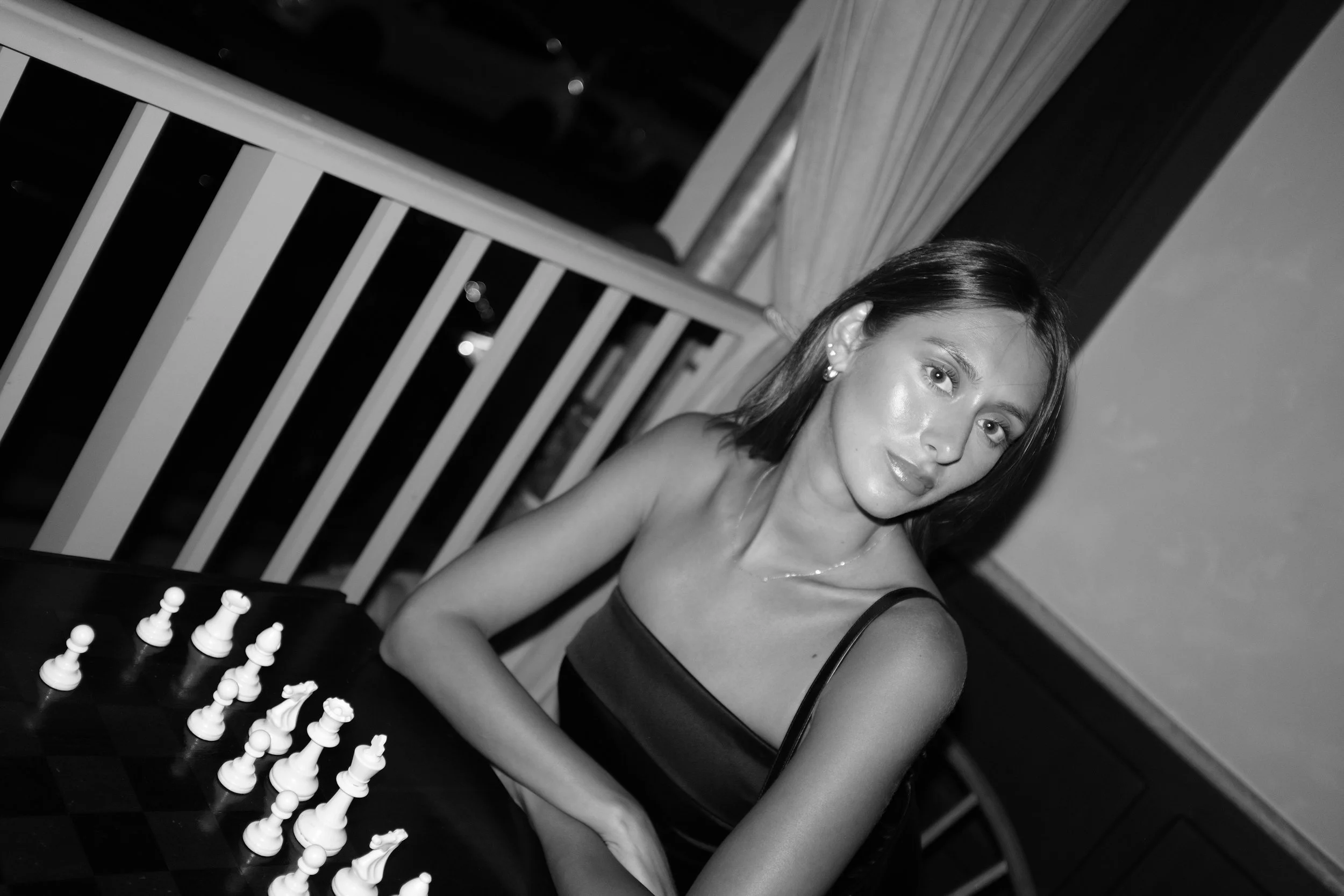 A mid-length black-and-white portrait of Katia Mueller sitting on a balcony in front of a chess set, looking directly at the camera.