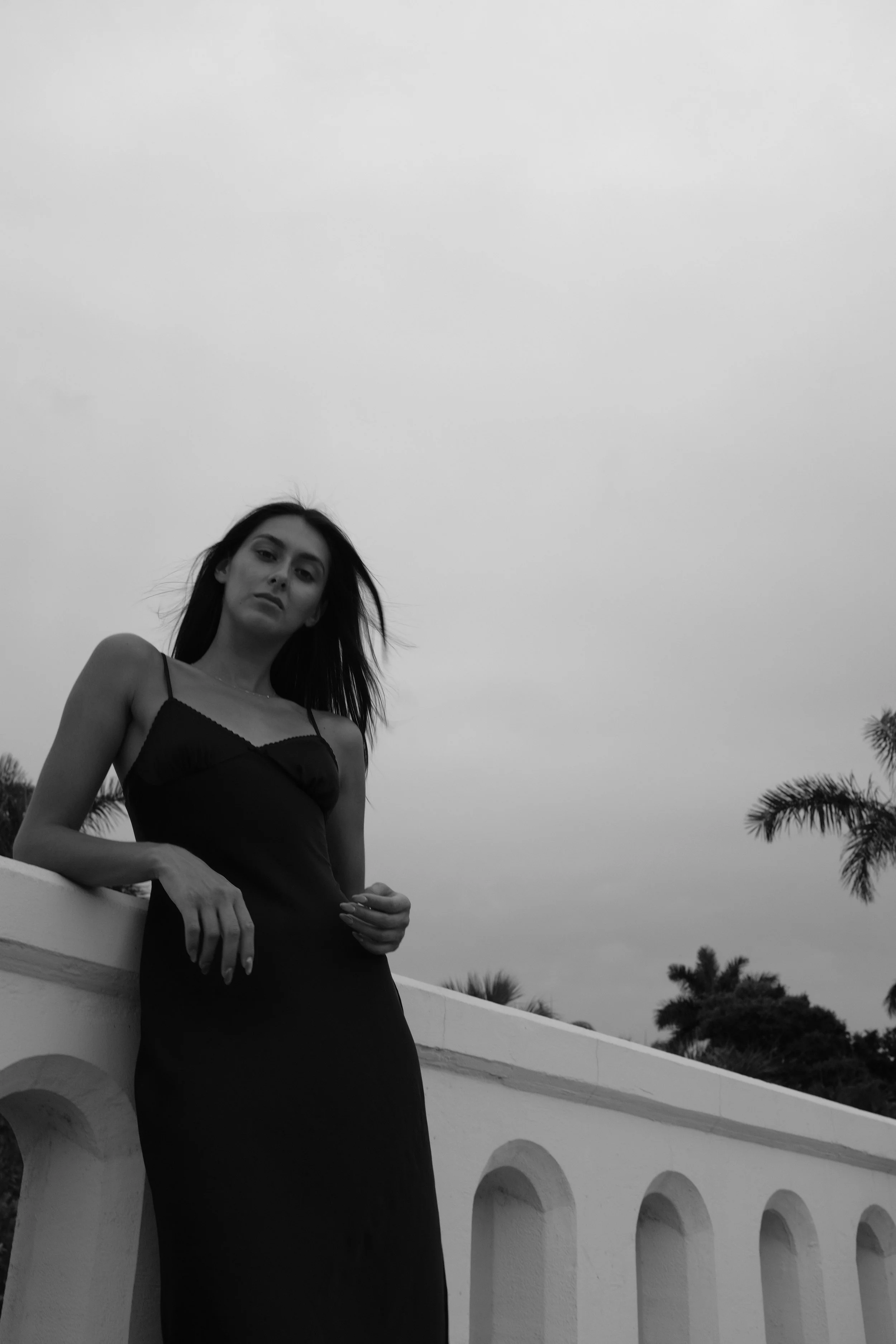 Black-and-white photo of a Katia Mueller wearing a long dark dress, standing on a curved outdoor balcony with palm trees in the background.