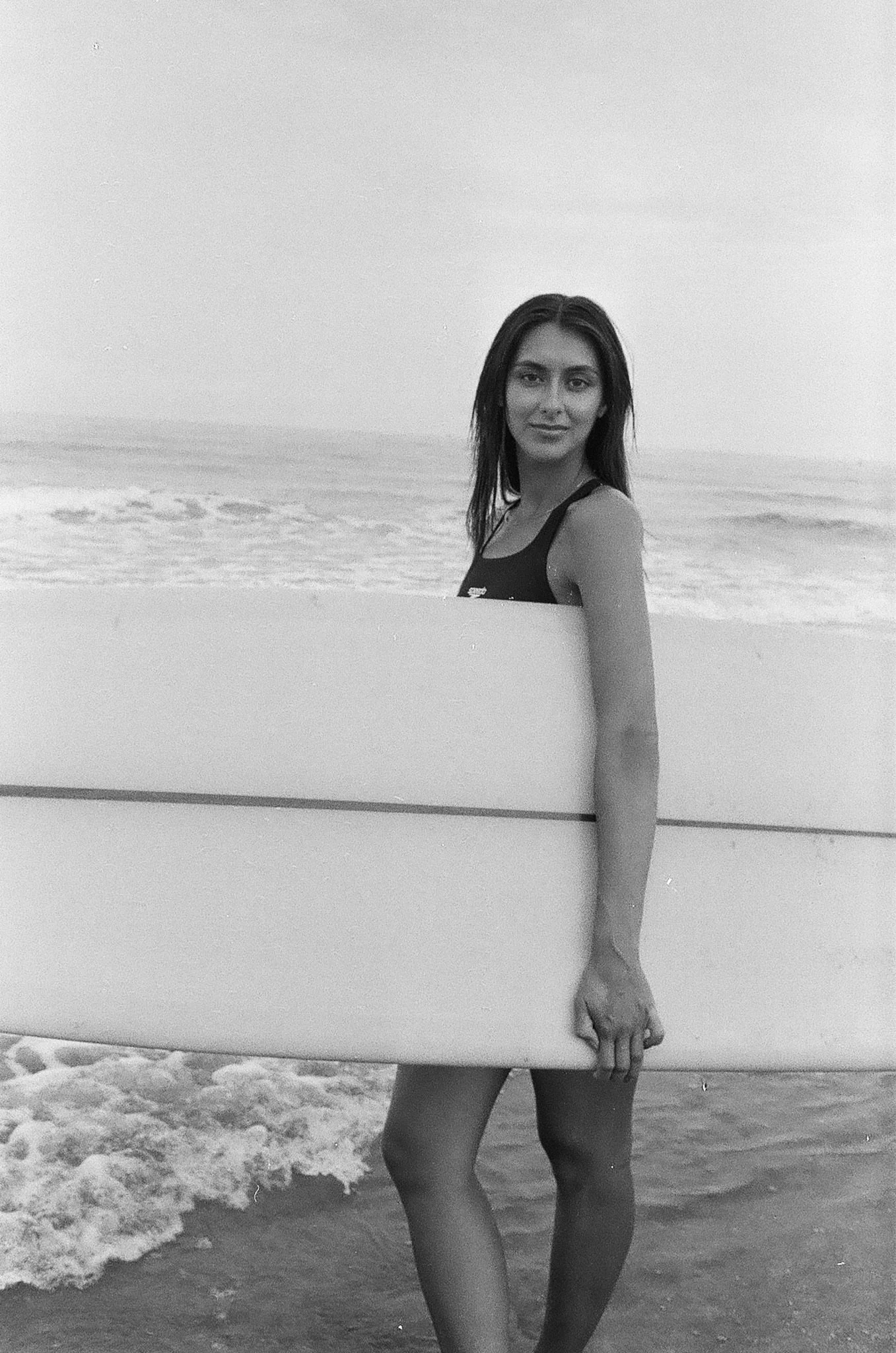 A three-quarters black-and-white portrait of Katia Mueller standing on the beach at the edge of the shore as she holds a surfboard, looking directly at the camera.