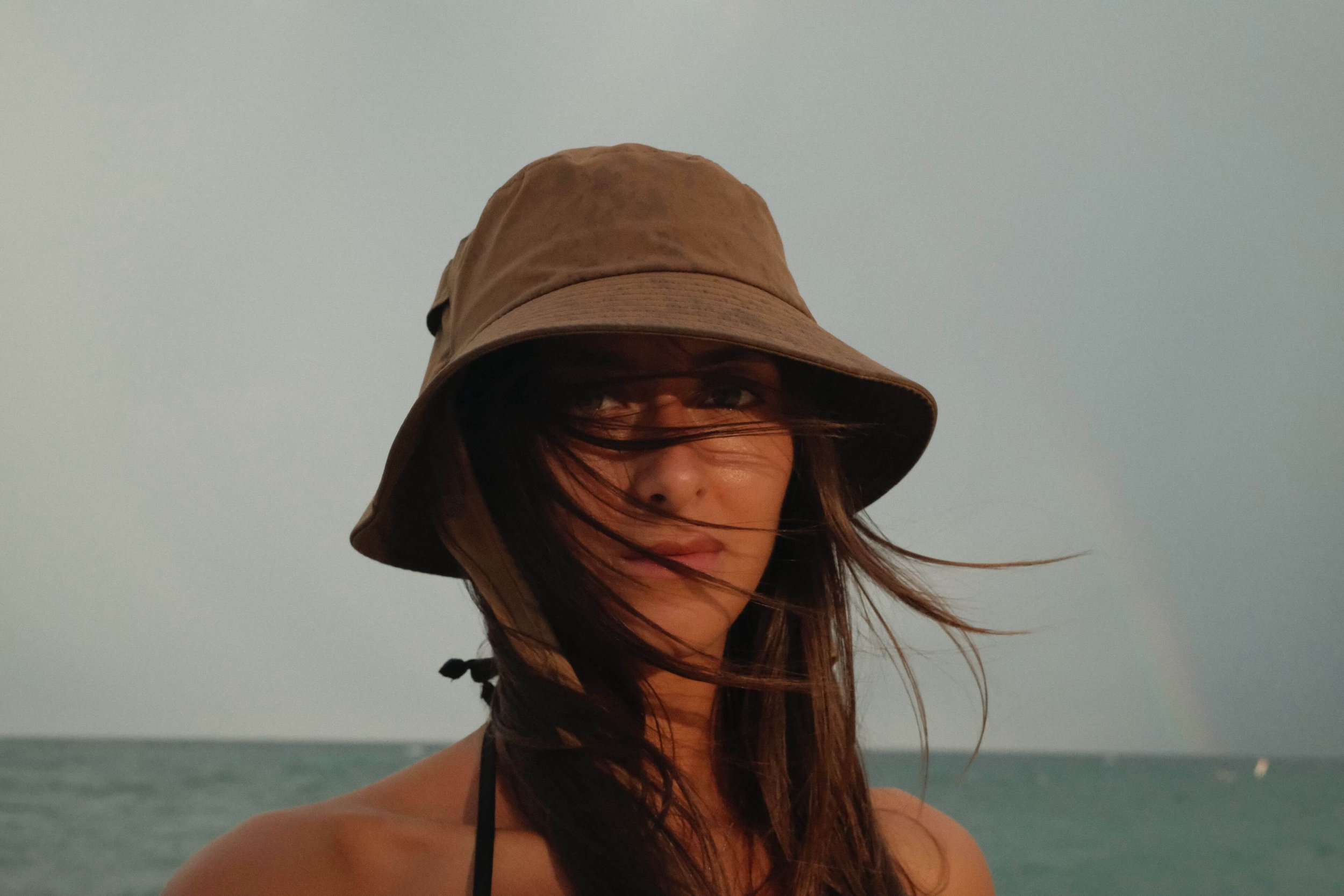 Katia Mueller wearing a brown bucket hat standing by the ocean, with wind blowing her hair across her face.