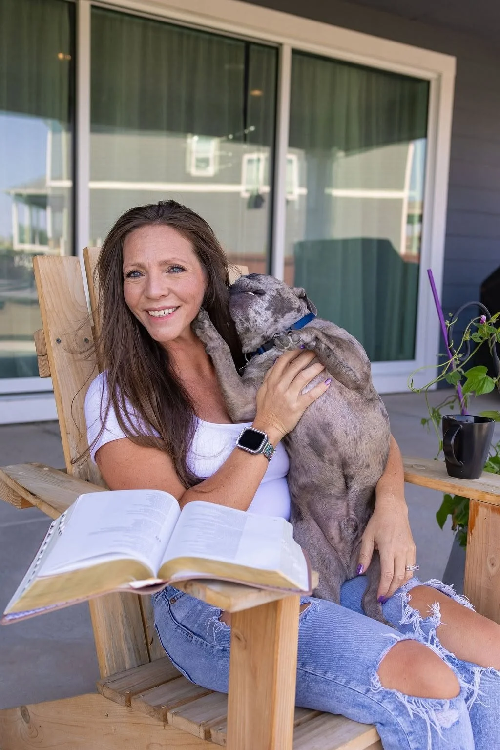 Woman with long brown hair smiling while holding a large gray dog on her lap, sitting on a wooden chair on a porch with a sliding glass door in the background.