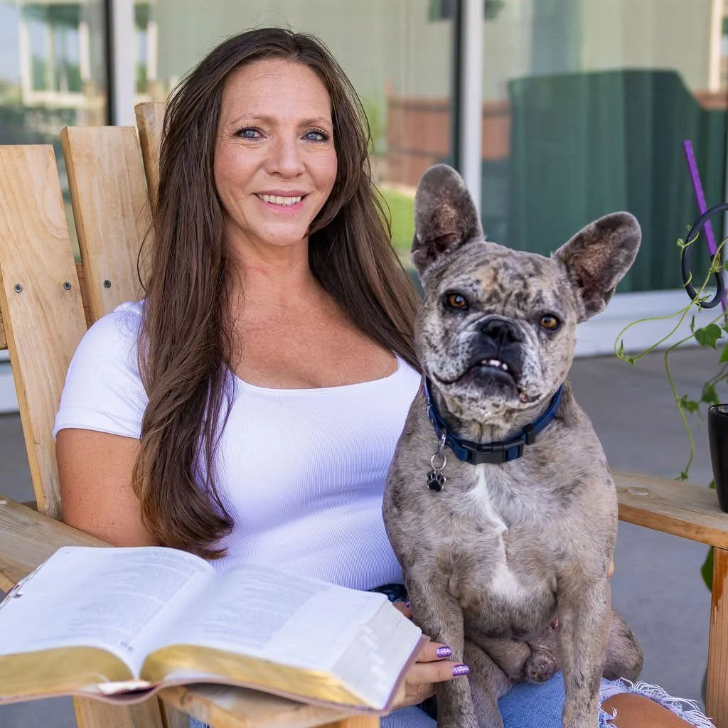 A woman sitting outdoors with a brick-colored French Bulldog puppy on her lap, holding an open book and smiling.