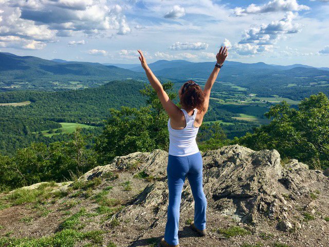 Lisa Hadlock standing on a rocky hilltop with her arms raised, overlooking a scenic landscape of green fields, trees, and distant mountains under a partly cloudy sky.