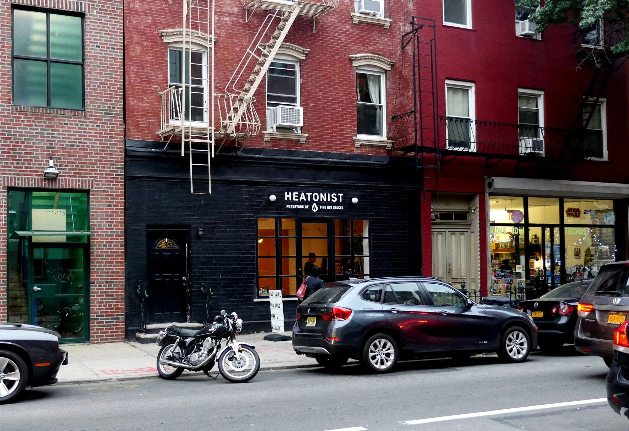 City street scene with parked cars, a motorcycle, a storefront called 'Heatonist' with a sign promoting hot sauces, and residential buildings with fire escapes.