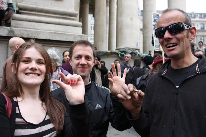 Three smiling people waving at the camera holding seed pills during an outdoor event in front of a building with columns.