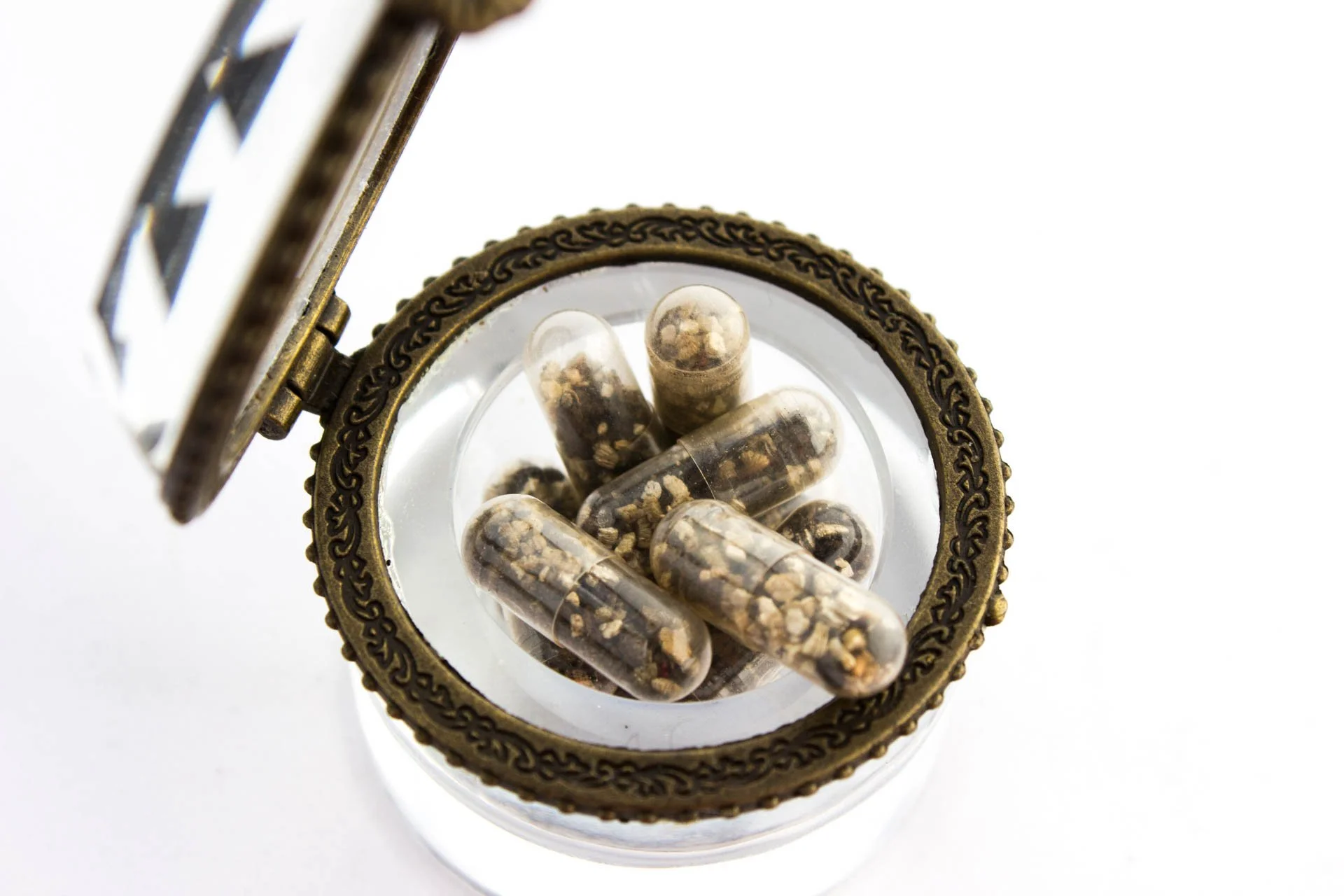 View of a small glass container with a decorative brass lid, containing capsules filled with dried herbs or plant material.