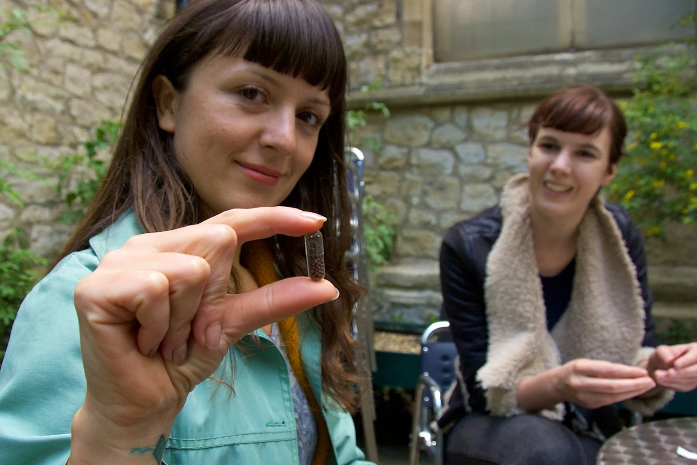 A woman holding a seed pill containing seeds, with another woman smiling in the background outside near a stone wall.