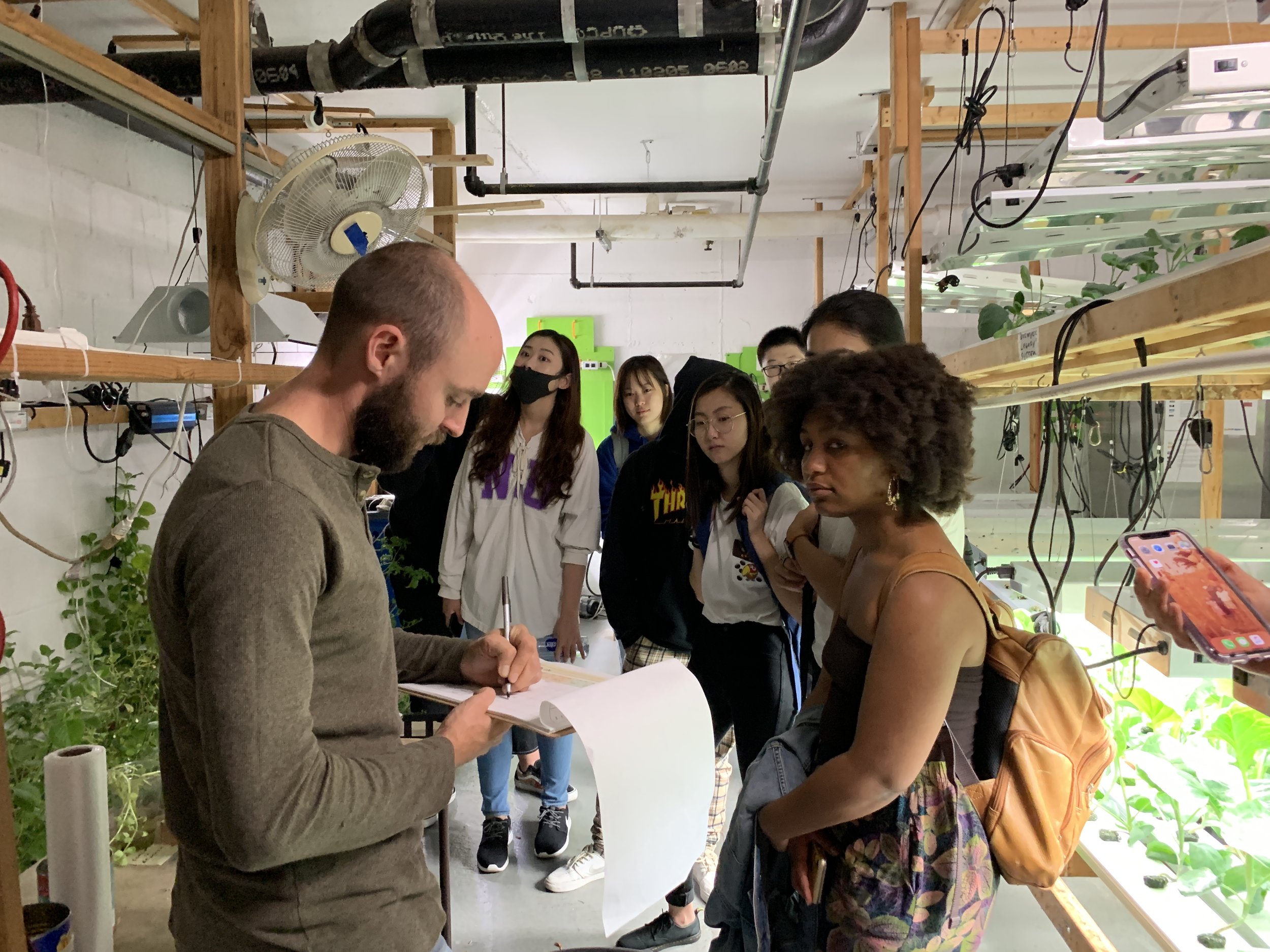 A man with a beard is writing in a notebook while a group of women and girls watch him, inside a plant-growing greenhouse with shelves of plants and grow lights.