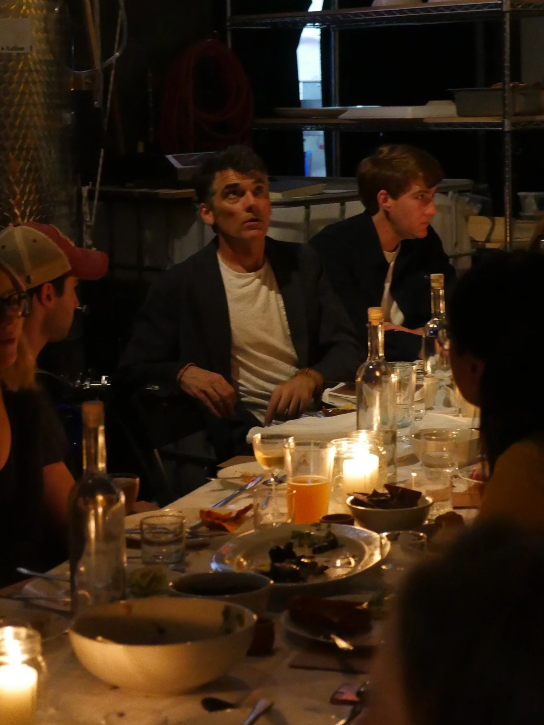 People gathered around a dining table in a dimly lit setting, with food, drinks, and a candle.