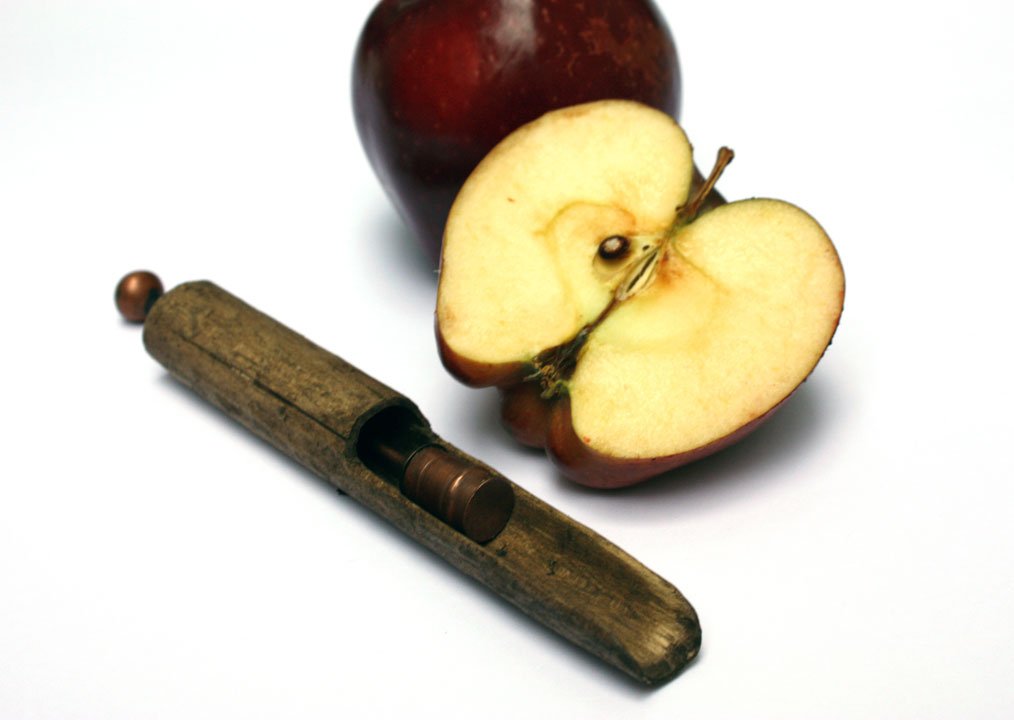 A halved red apple with a stem, a whole red apple, and a small wooden object with a sliding mechanism on a white surface.