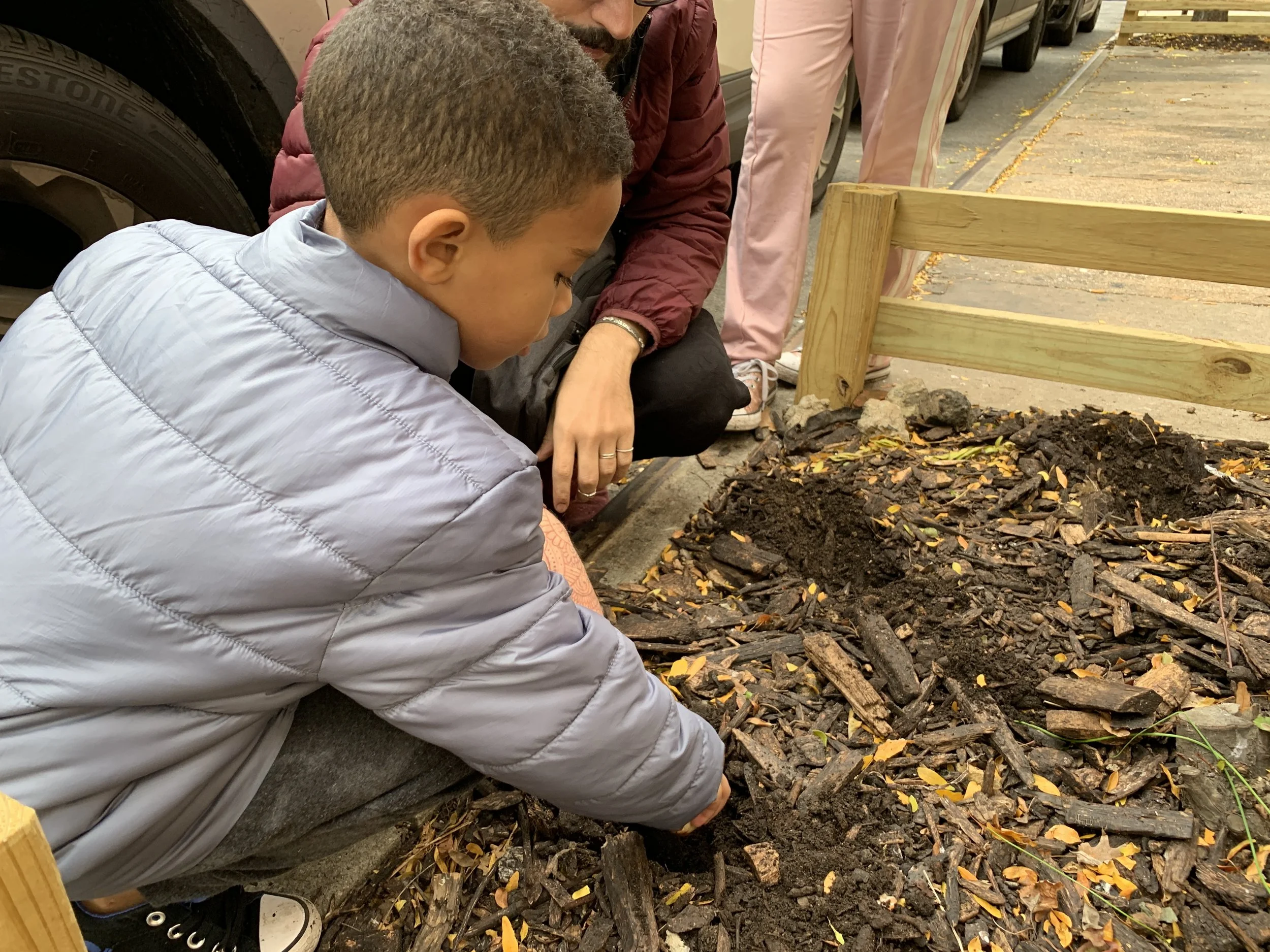 A young boy planting seeds or small plants into soil alongside an adult woman, with wood mulch and a wooden fence nearby.
