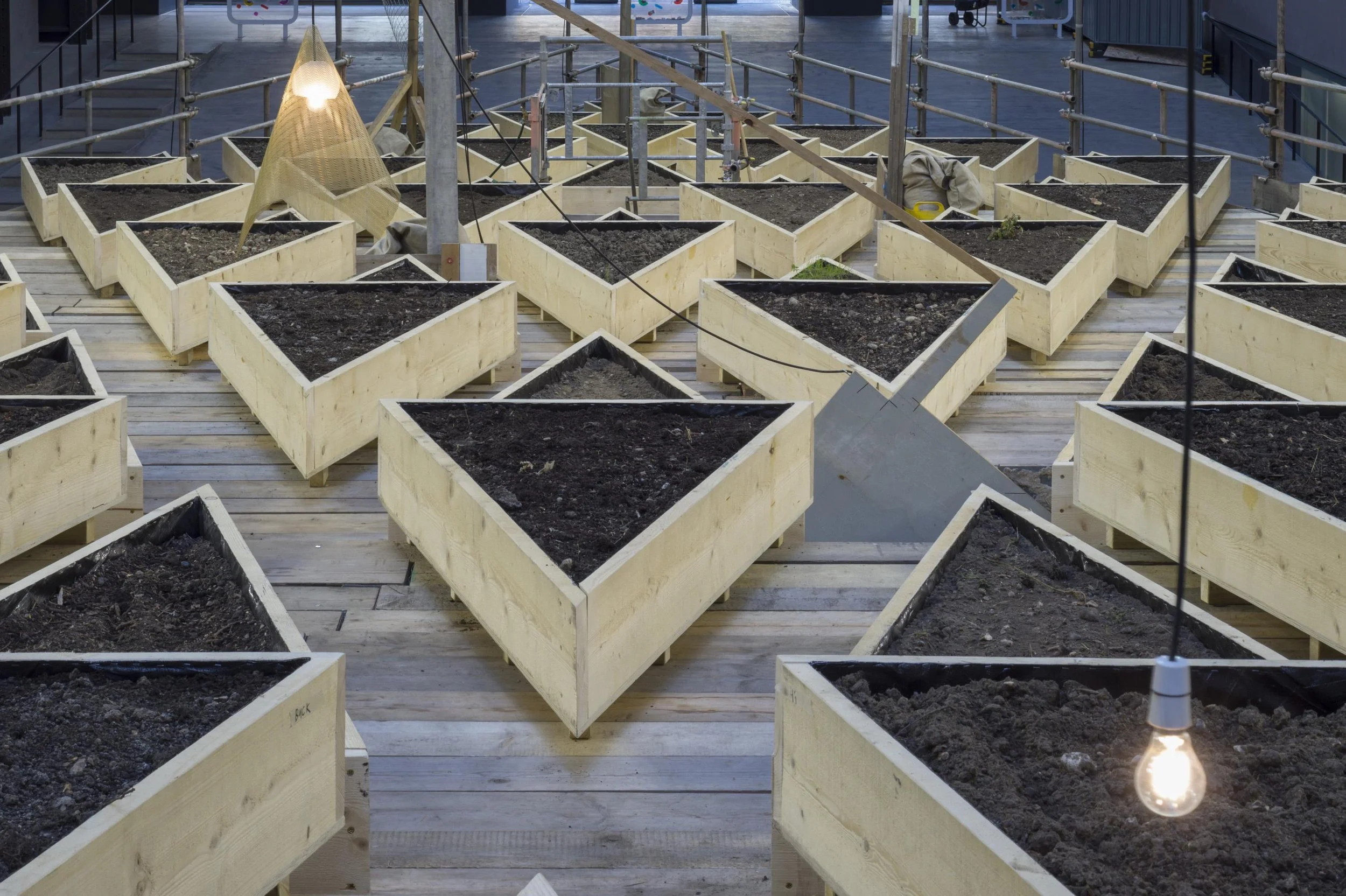 Construction site with multiple wooden planters filled with soil. The planters are arranged on a wooden deck with a safety railing around the area. Temporary lighting fixtures hang above, and there are some construction materials and equipment around.