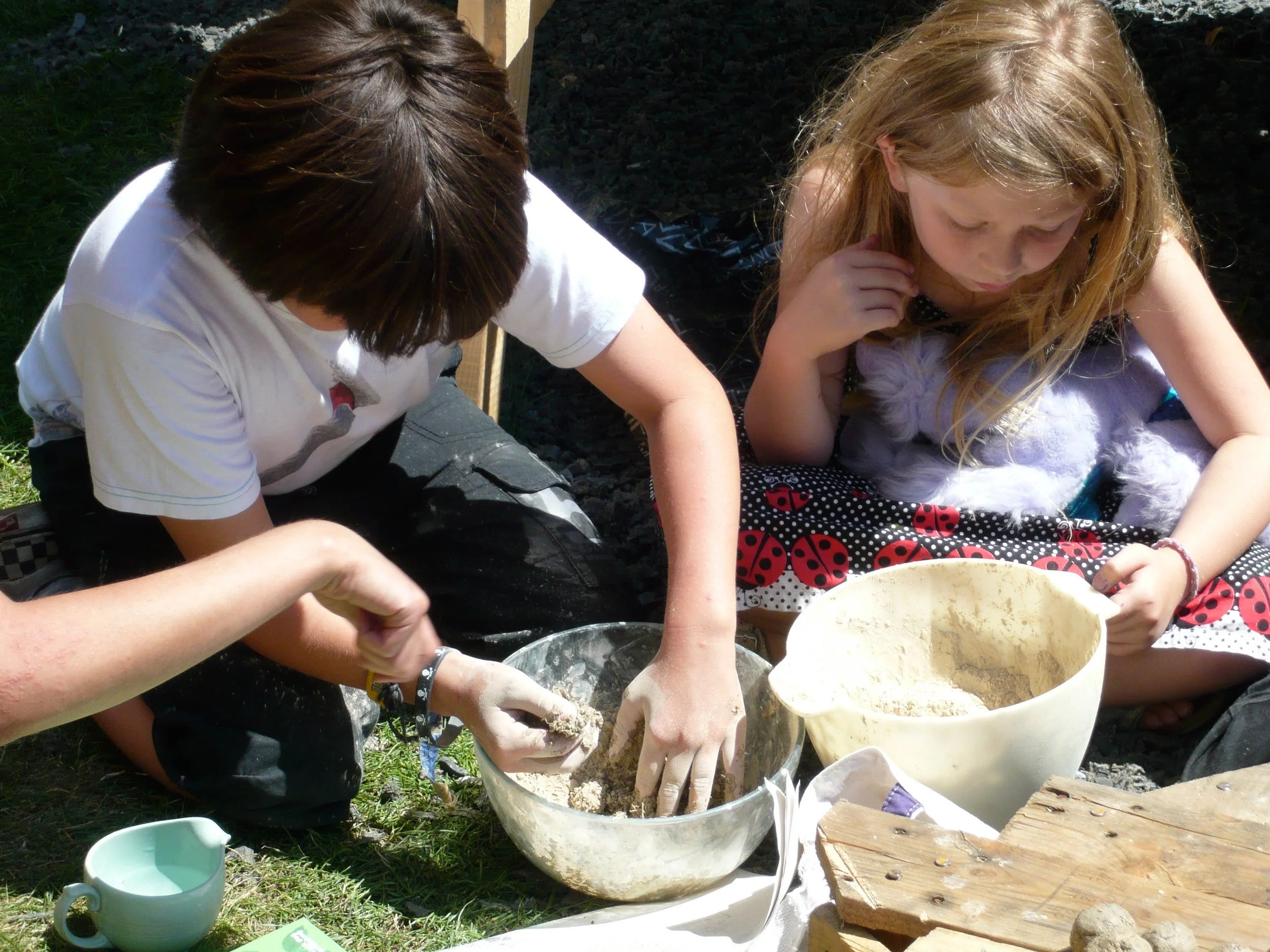 Two children sitting outdoors, mixing something in a glass bowl with their hands, with a large beige bowl nearby. One child has long blonde hair and a ladybug dress, the other has dark hair and a white t-shirt. There are various objects including a small blue mug and a wooden block around them.