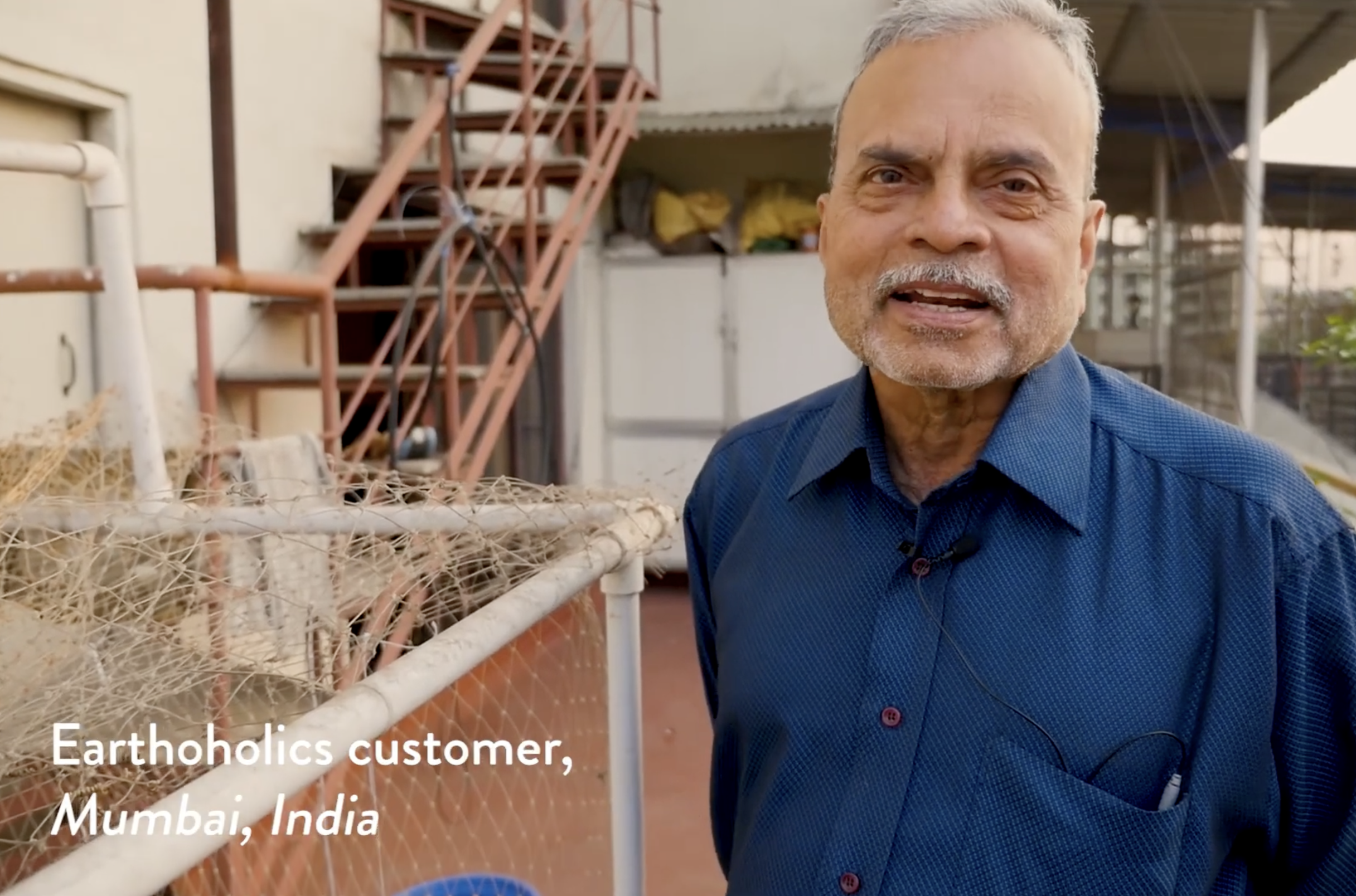 An elderly man with gray hair and a beard, wearing a blue shirt, standing on a balcony with a metal staircase and a large cage or enclosure with netting in Mumbai, India.