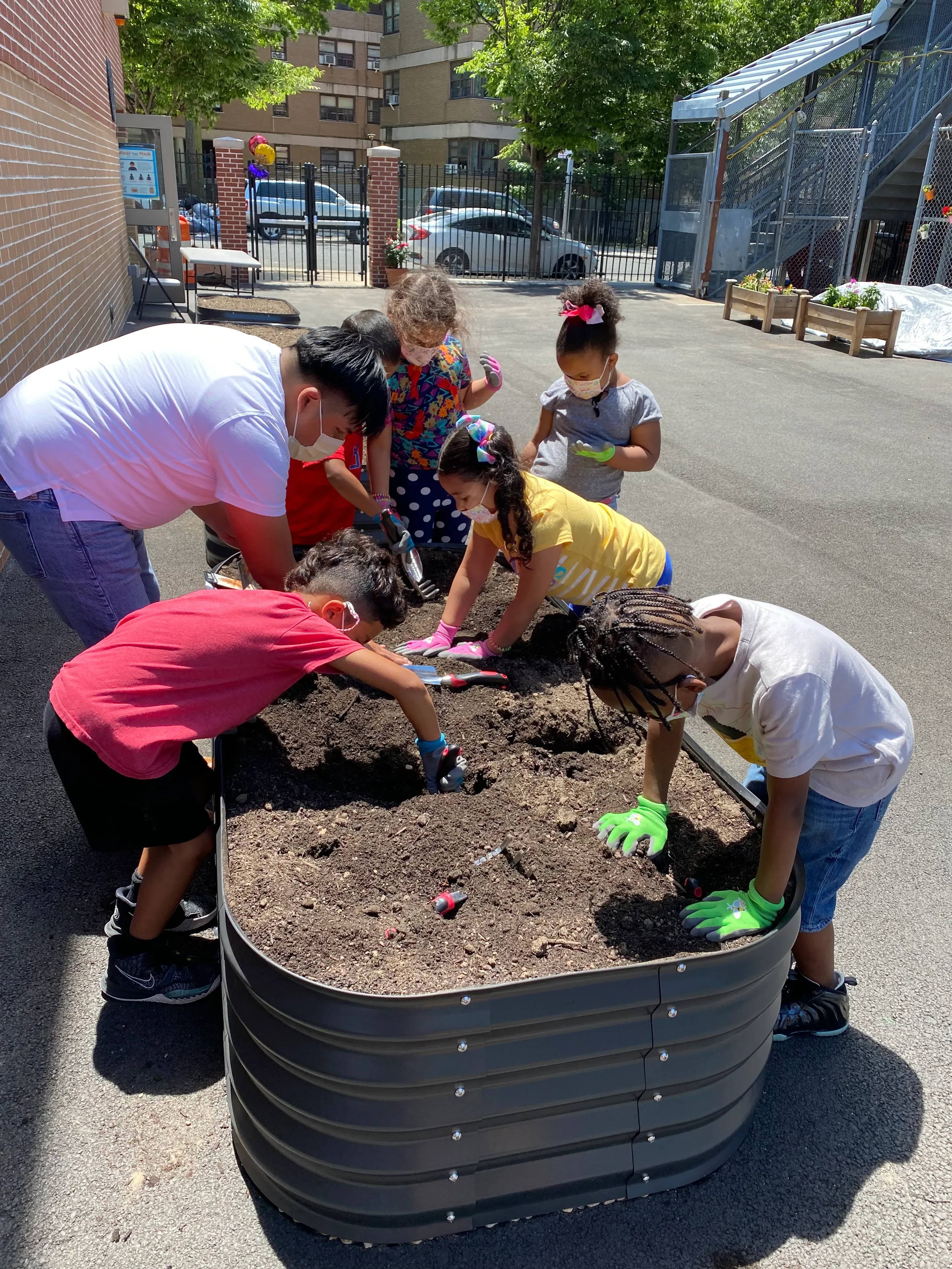 Children and an adult planting flowers in a large outdoor garden bed, all wearing masks and gloves, on a sunny day.