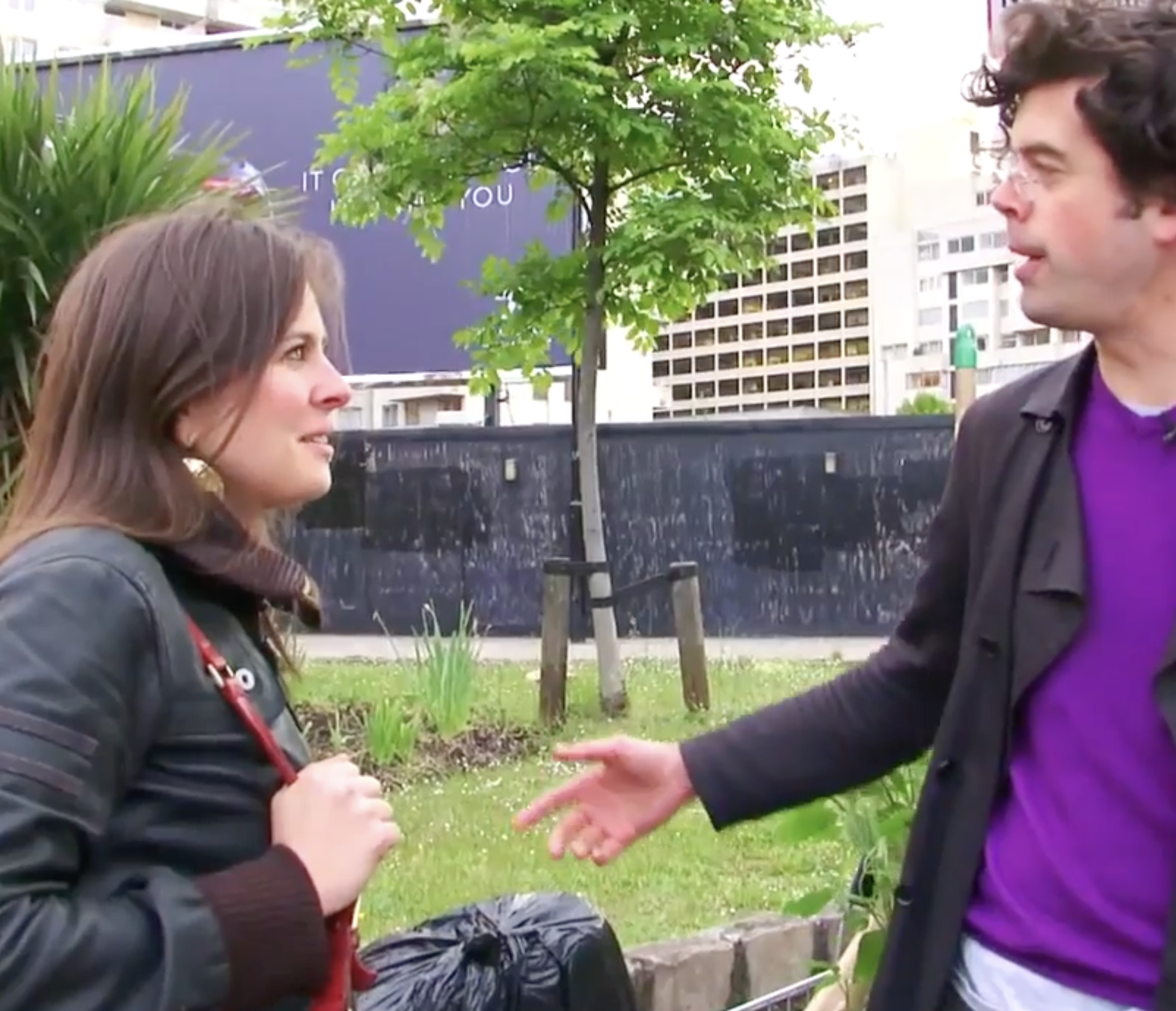 A woman and a man having a conversation outdoors in a city park, with buildings and trees in the background.
