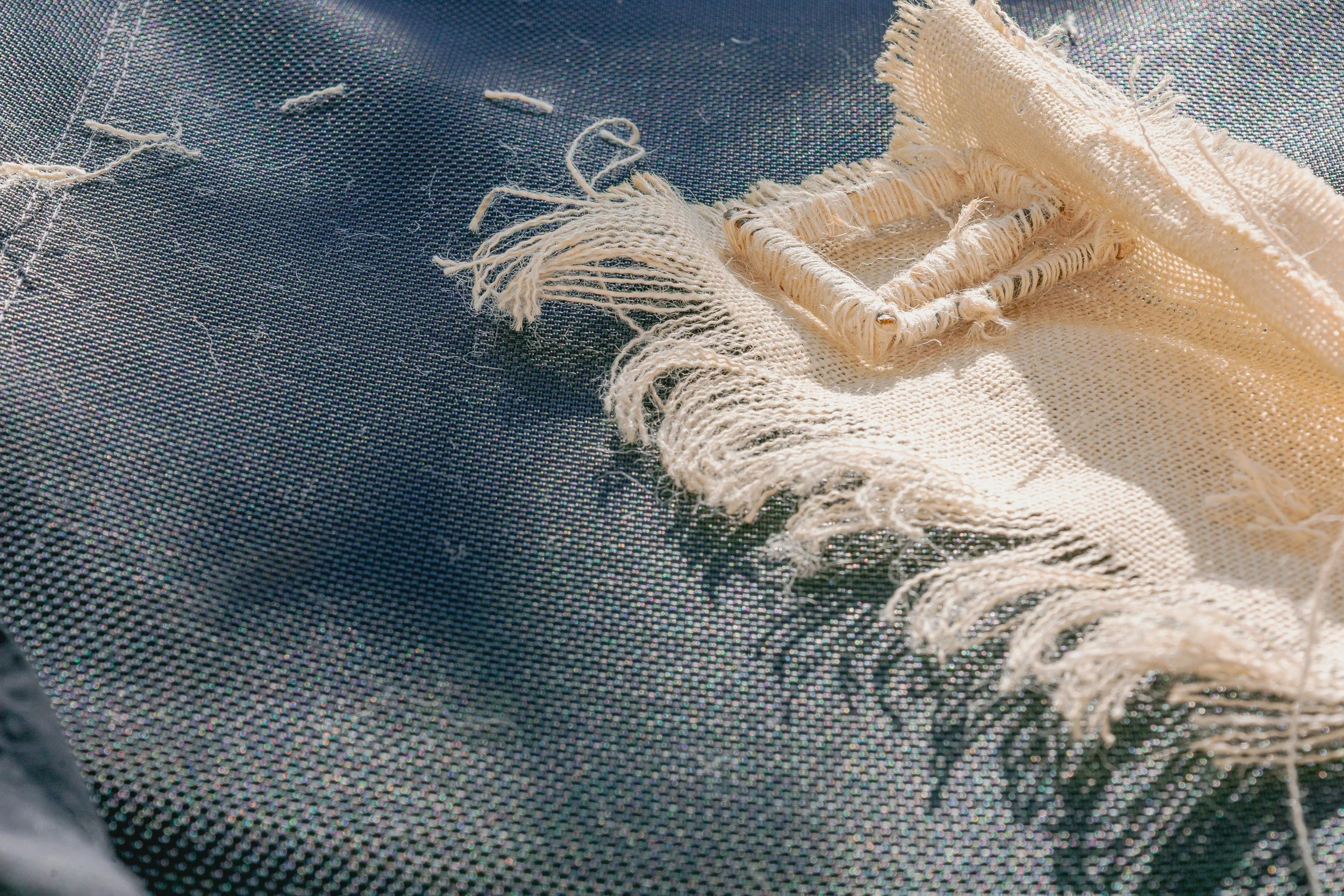 Close-up of a frayed, cream-colored textured fabric with a square woven label, resting on dark perforated material.