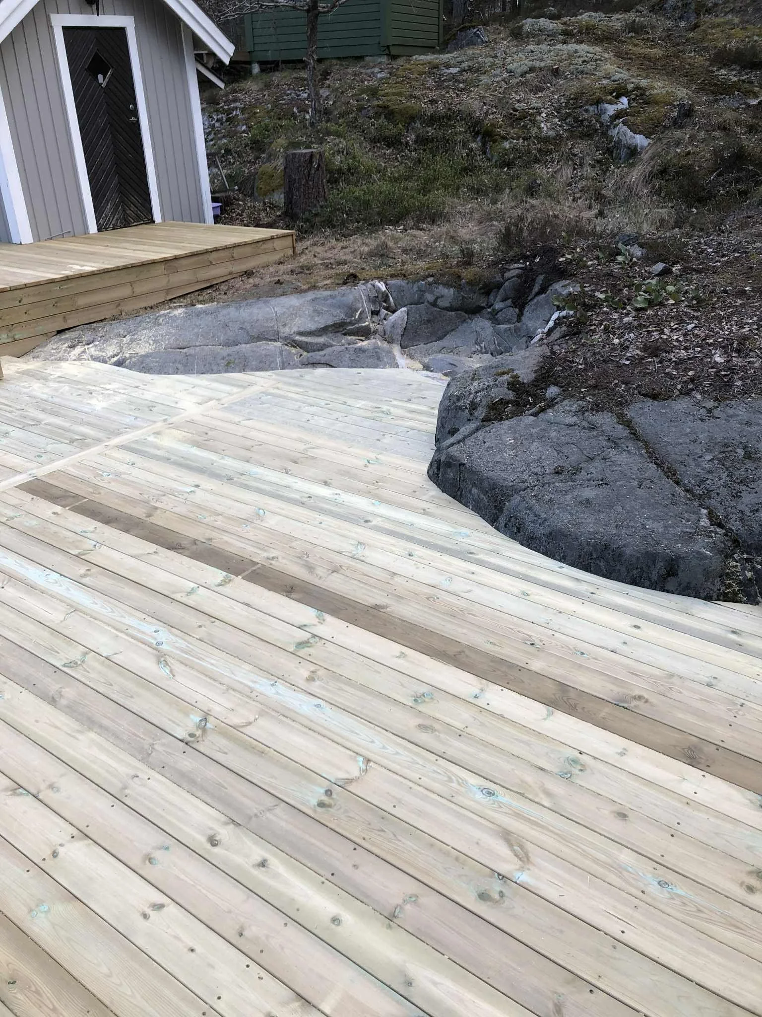 Nybyggd trädäckad uteplats med trappsteg intill en liten stuga, omgiven av steniga och naturliga markområden.