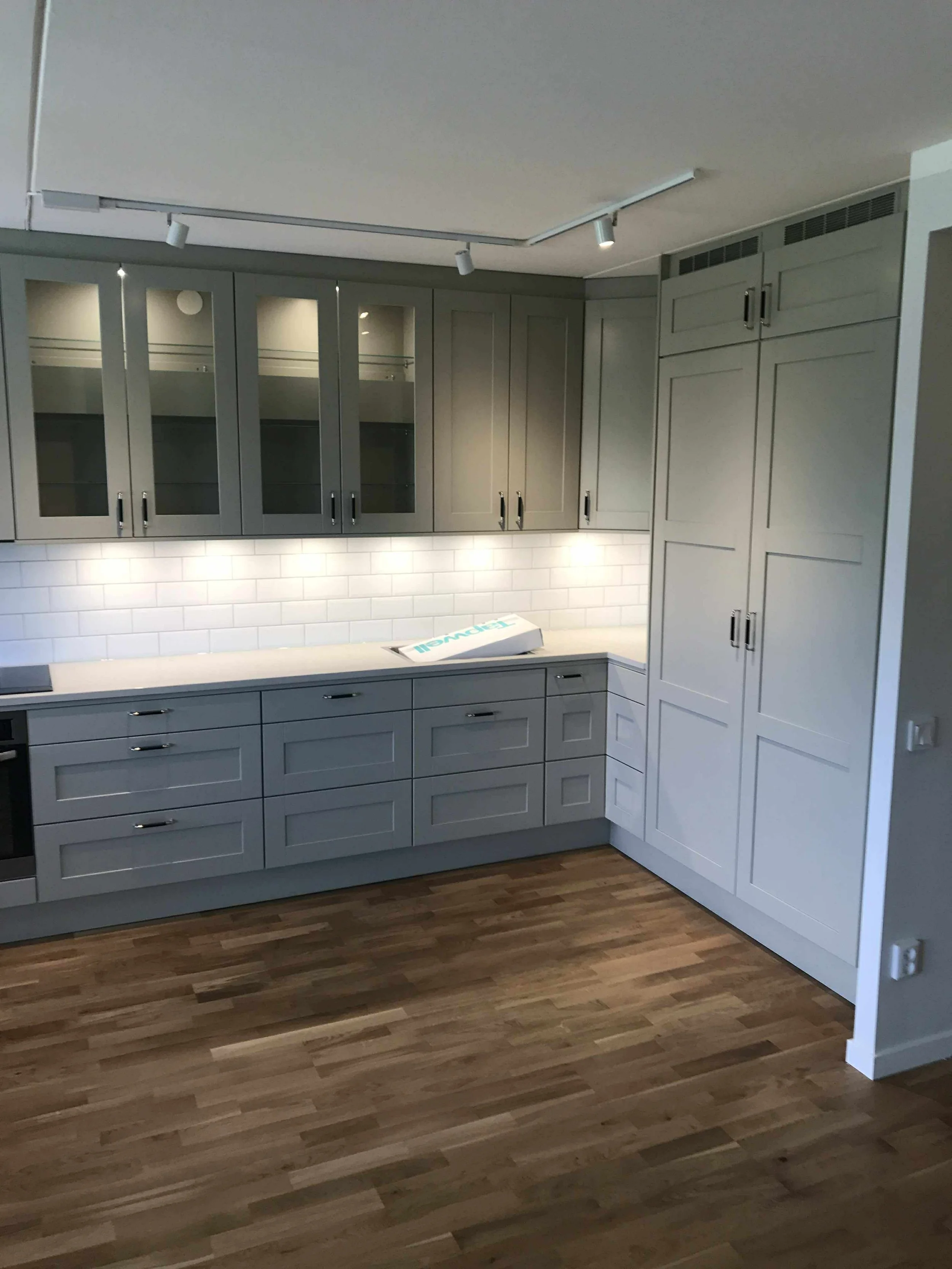 En modern köksinredning med grå skåp, vita kakelväggar och trägolv.