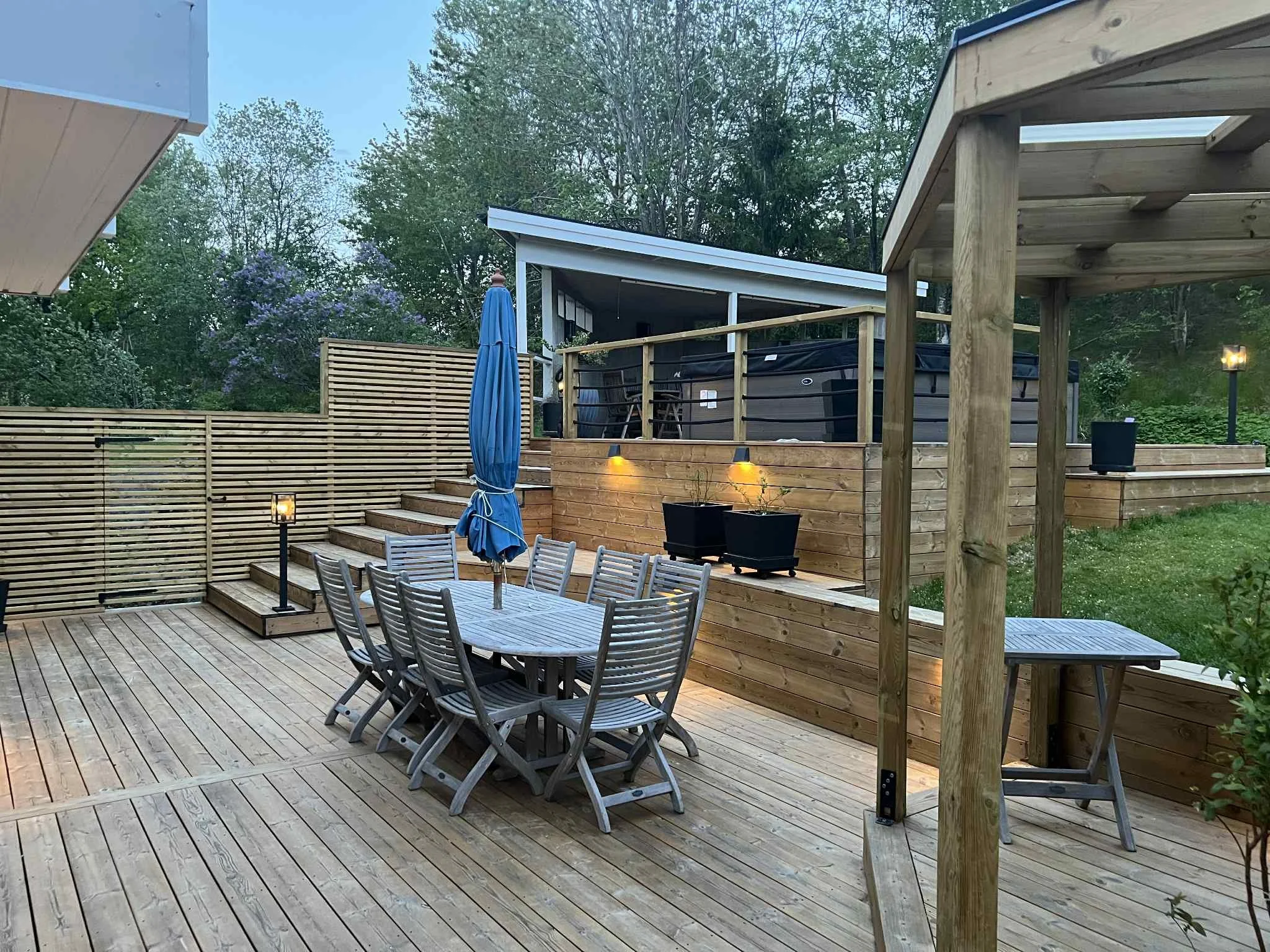 Uteplats med ett runt bord och flera stolar, ett parasoll, trappsteg, ett jacuzzi, växter och träd i bakgrunden, och utomhusbelysning.