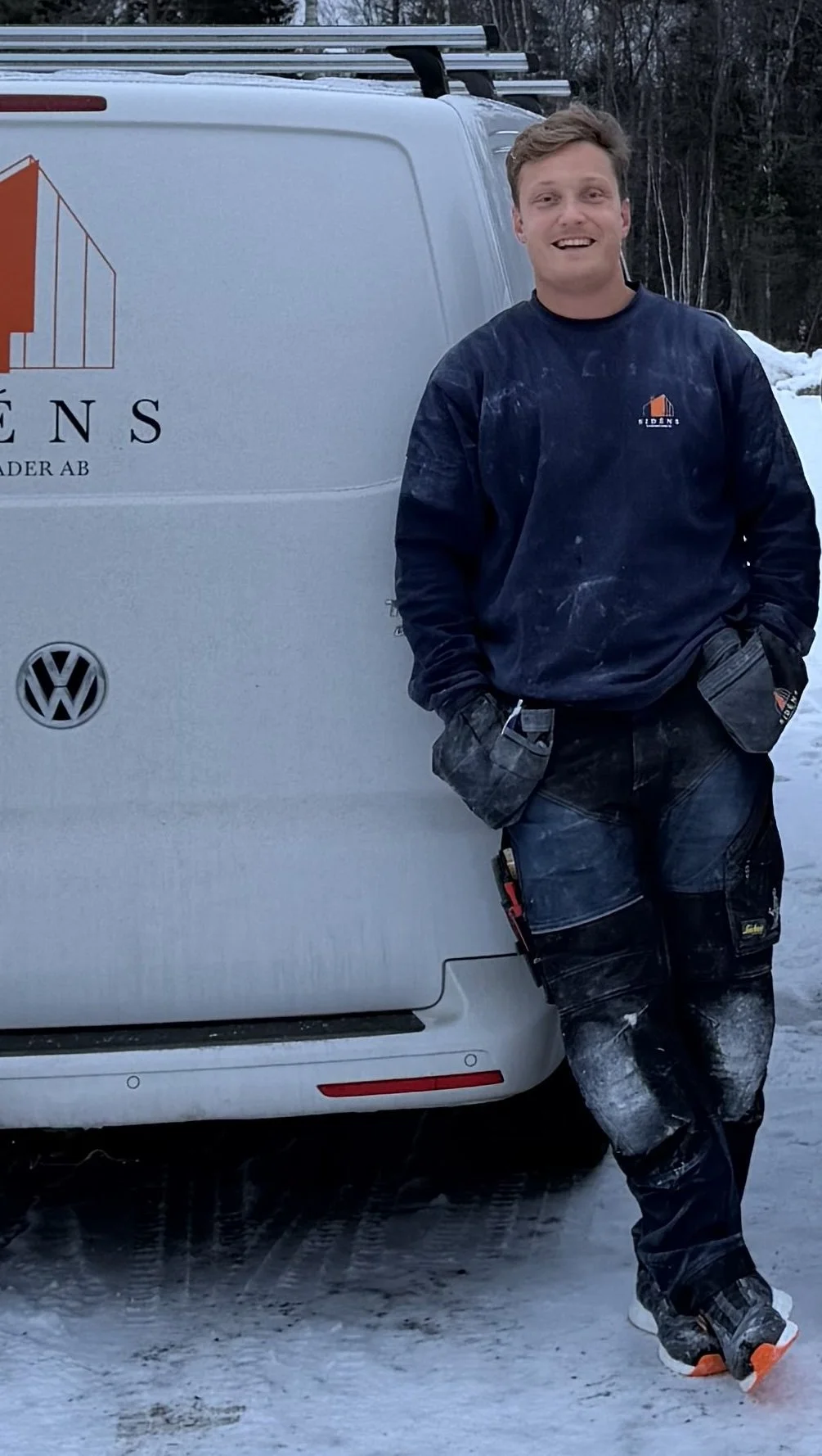 Linus står lutad mot en vit företagstransportbil med företagsnamnet Sidens Entreprenader AB på sidan, klädd i arbetskläder, i en snöig utomhusmiljö.