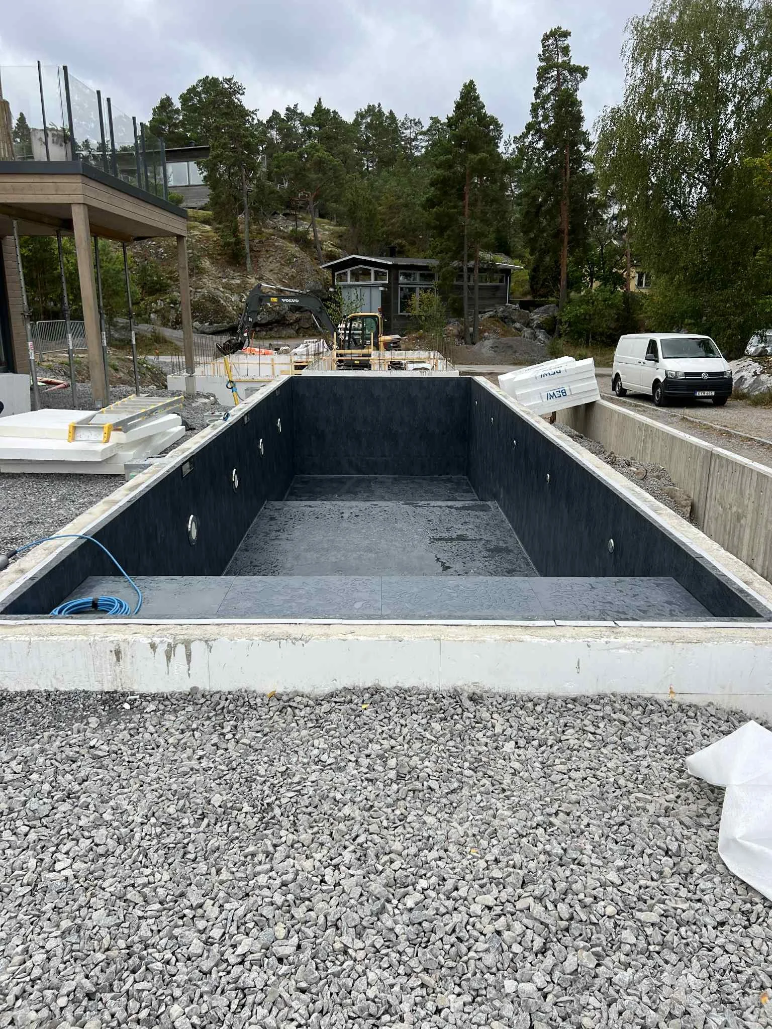 En tom pool under konstruktion, omgiven av byggmaterial och utrustning, i ett område med stenar och träd.