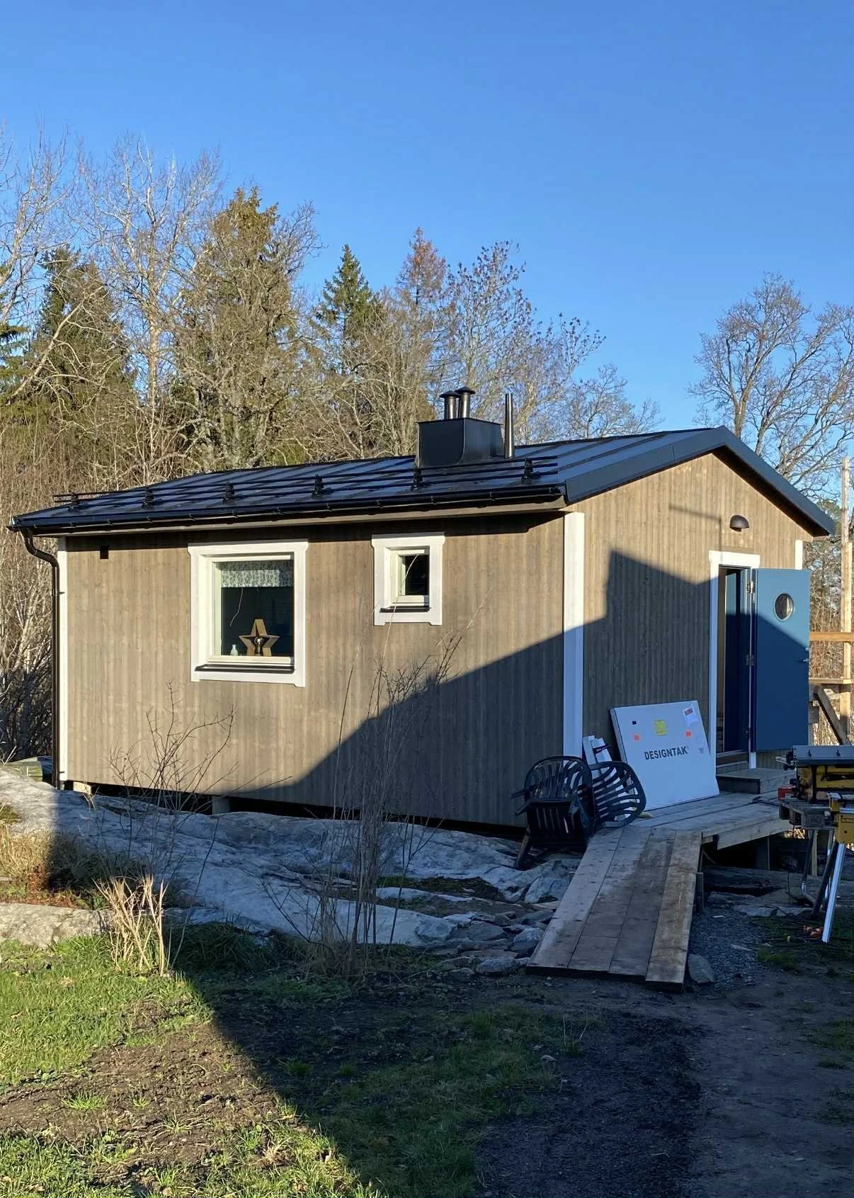 Ett nybyggt litet hus med träfasad och tak, som fortfarande är under konstruktion, omgiven av träd och en trädgård.