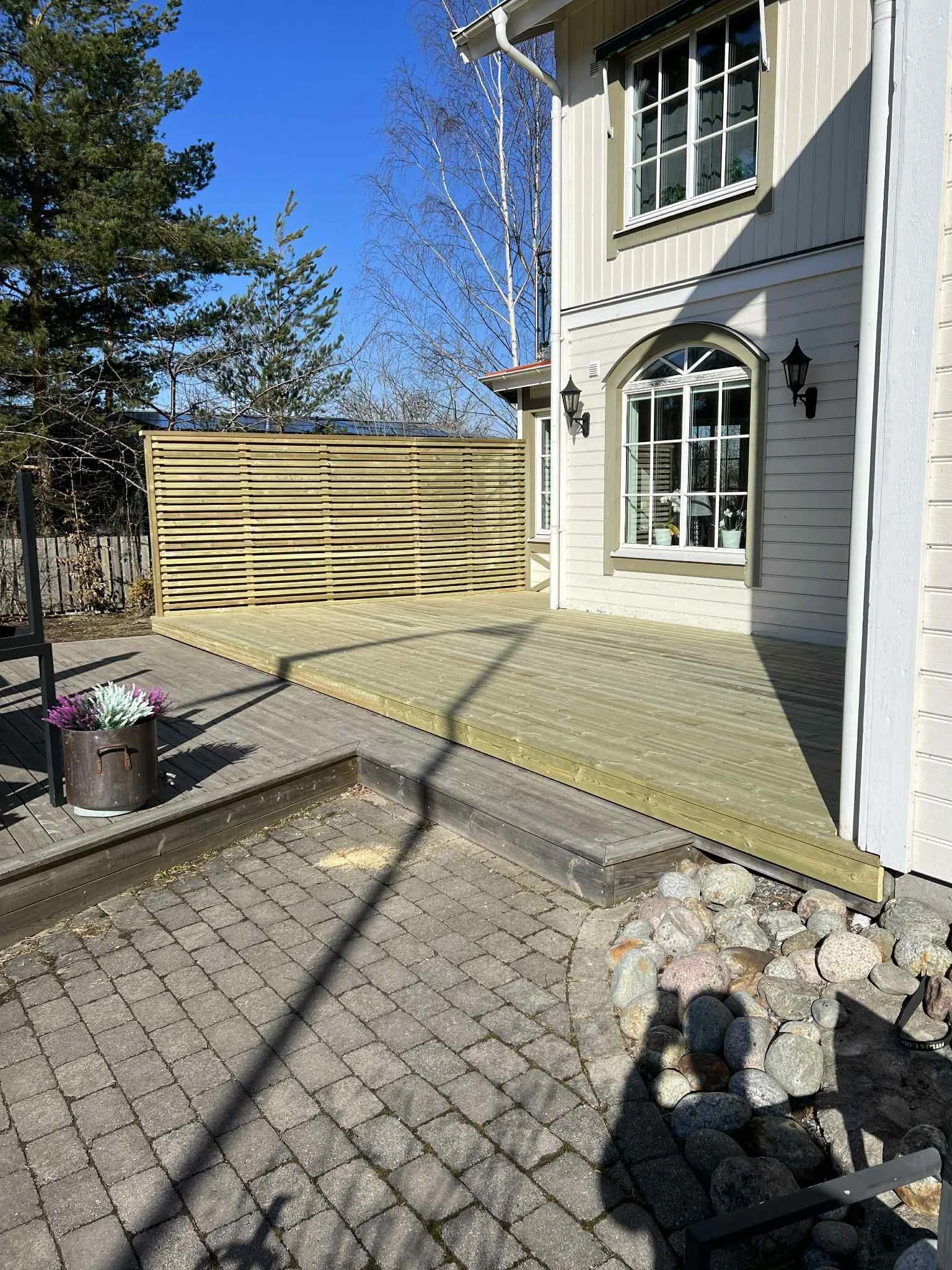 En utomhusveranda av trä framför ett vitt hus med stora fönster och två vägglampor, omgiven av en stenlagd gård och några träd.