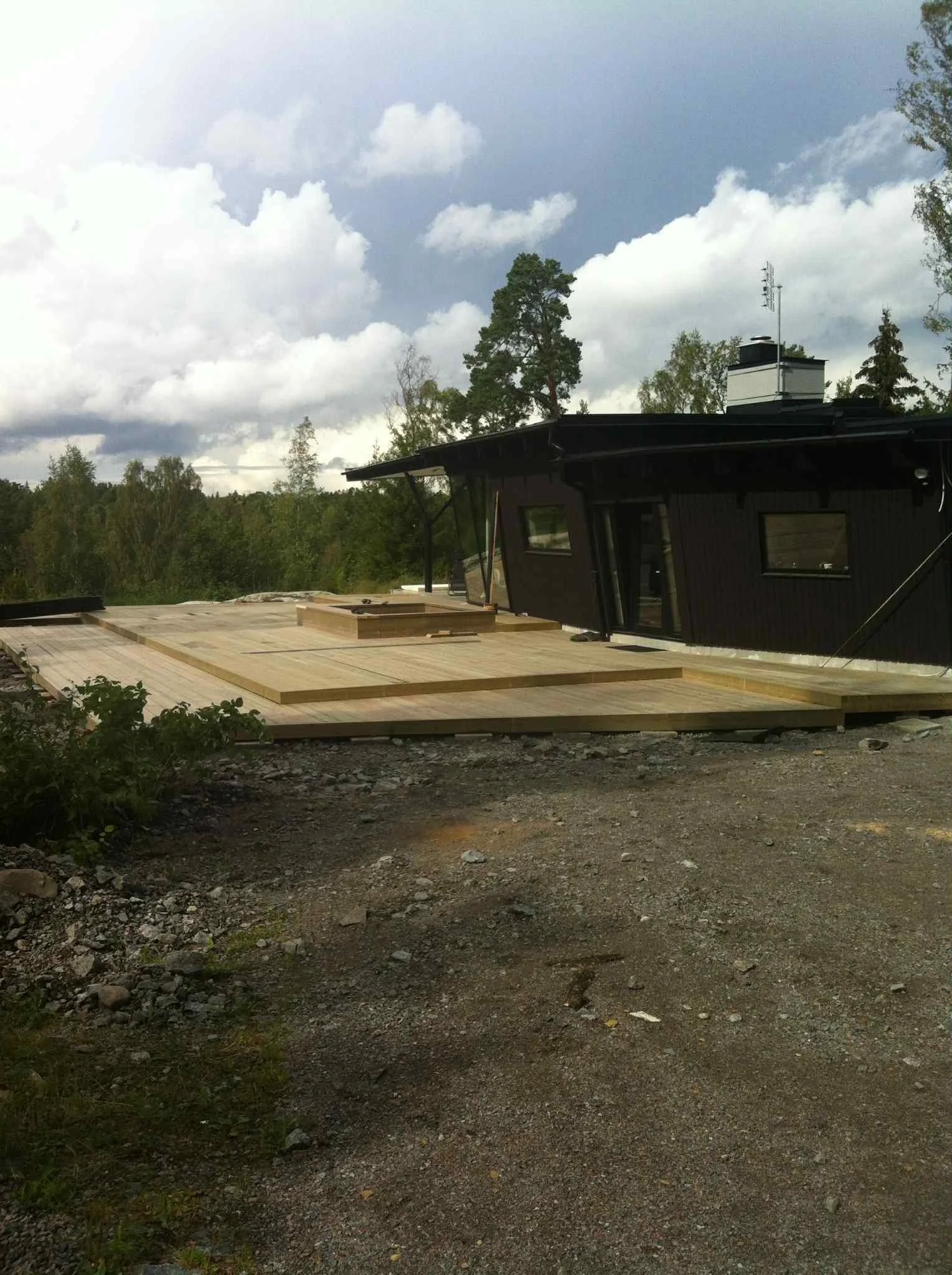 Nybyggd trädäckad uteplats framför ett hus, omgiven av skog och träd, under en molnig himmel.