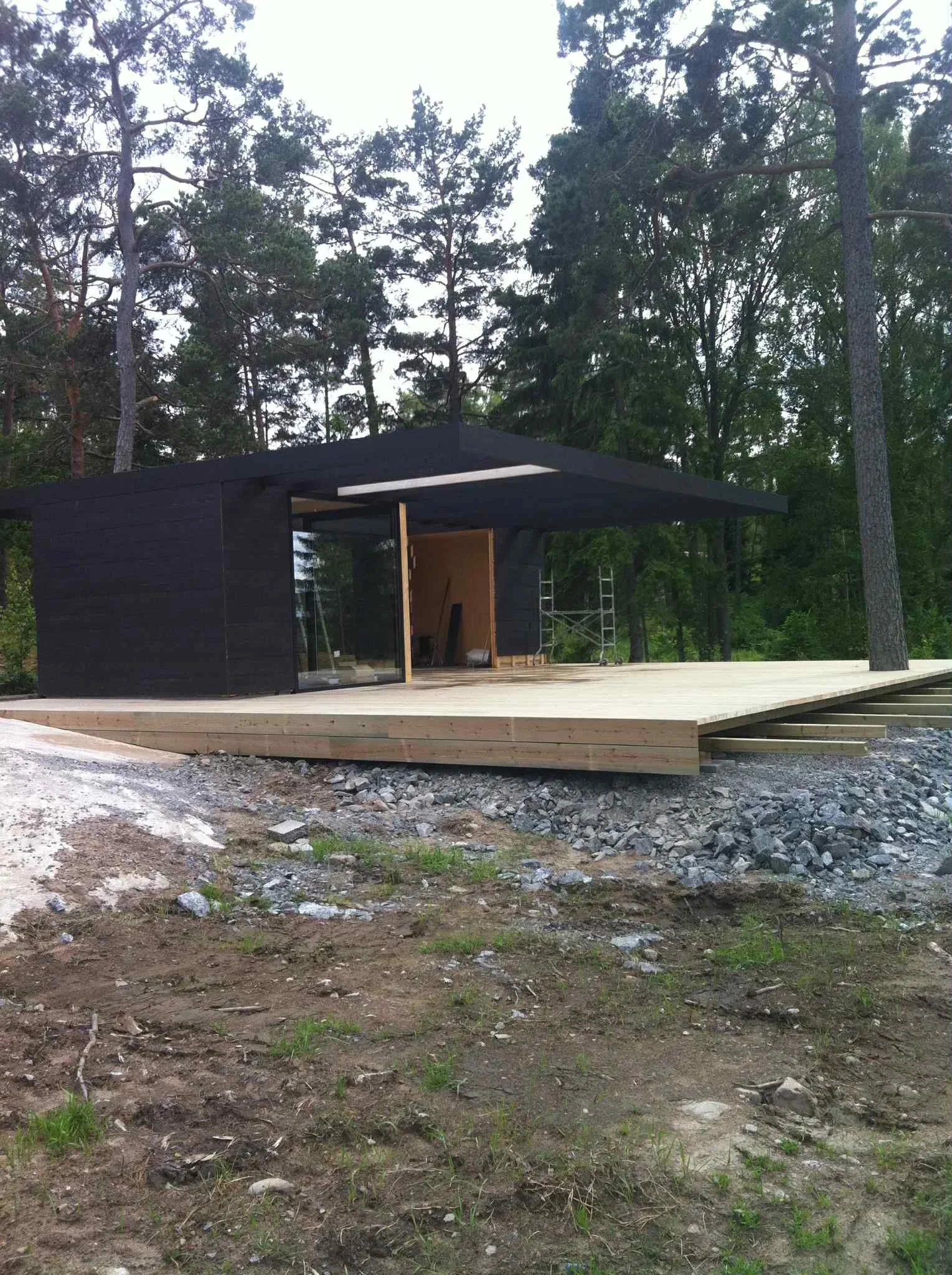 Nybyggt hus på en tränivilning, omgiven av tallträd, med trädäckad terrass under byggnation.