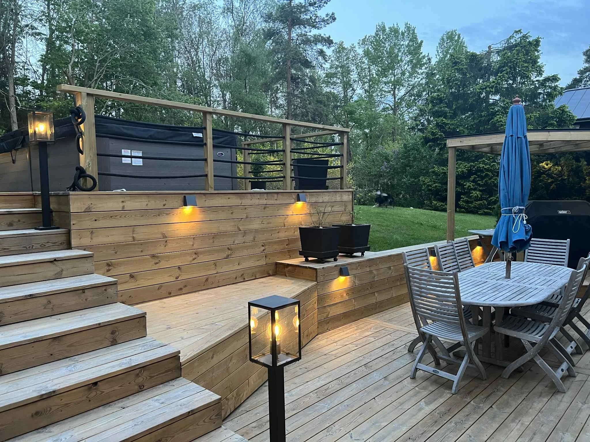 En trädäckad uteplats med trappsteg och en uteplats möblerad med ett runt bord, flera stolar och ett solskydd. Det finns även ljusstyl i form av vägglampor, ett golvljus och ett lyktglas. Bakom den används ett avgränsat skogområde med träd och buskar.