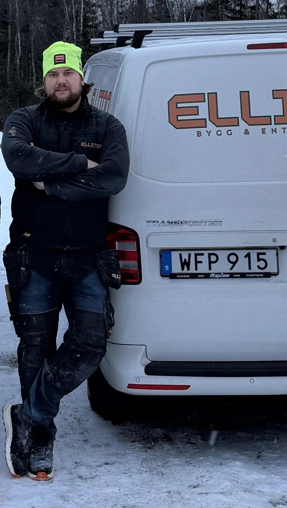 Vincent står bredvid en vit lastbil med företagsnamnet Elliton Bygg & Entreprenad på sidan, han är klädd i svart arbetskläder och en neongrön mössa, utomhus på snöig mark.