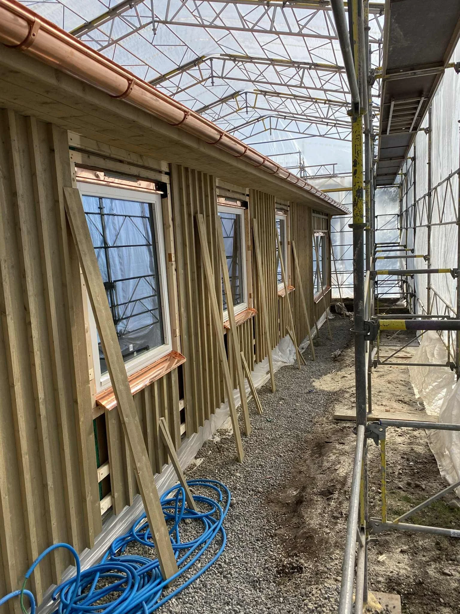 Byggarbetsplats med fönster och träpanel på ett hus under konstruktion, omgiven av byggställningar och skyddsnät.