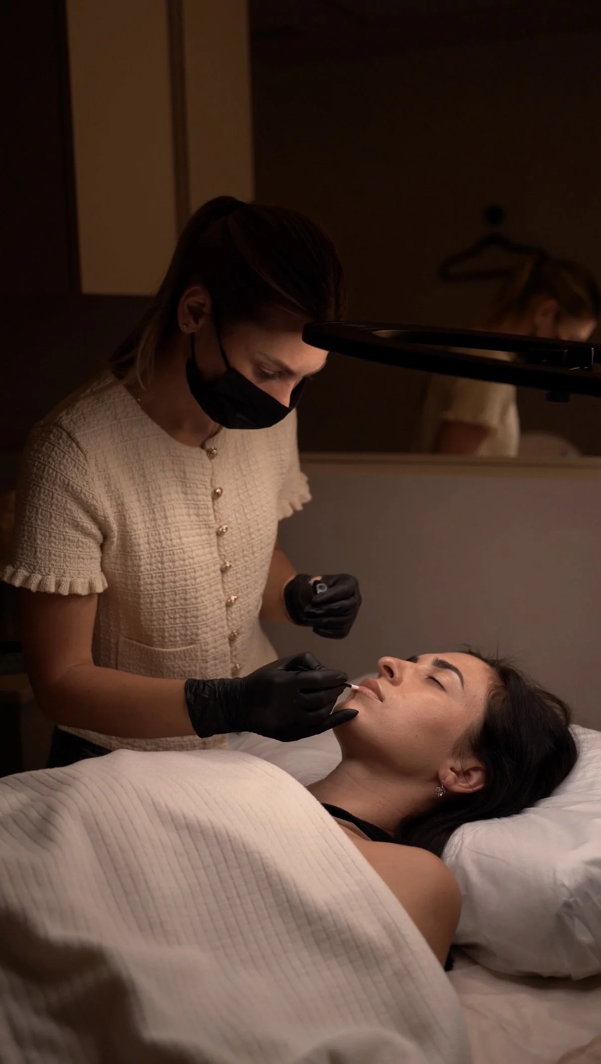 A woman lying on a bed receiving a cosmetic treatment from a professional wearing gloves and a face mask in a dimly lit room.