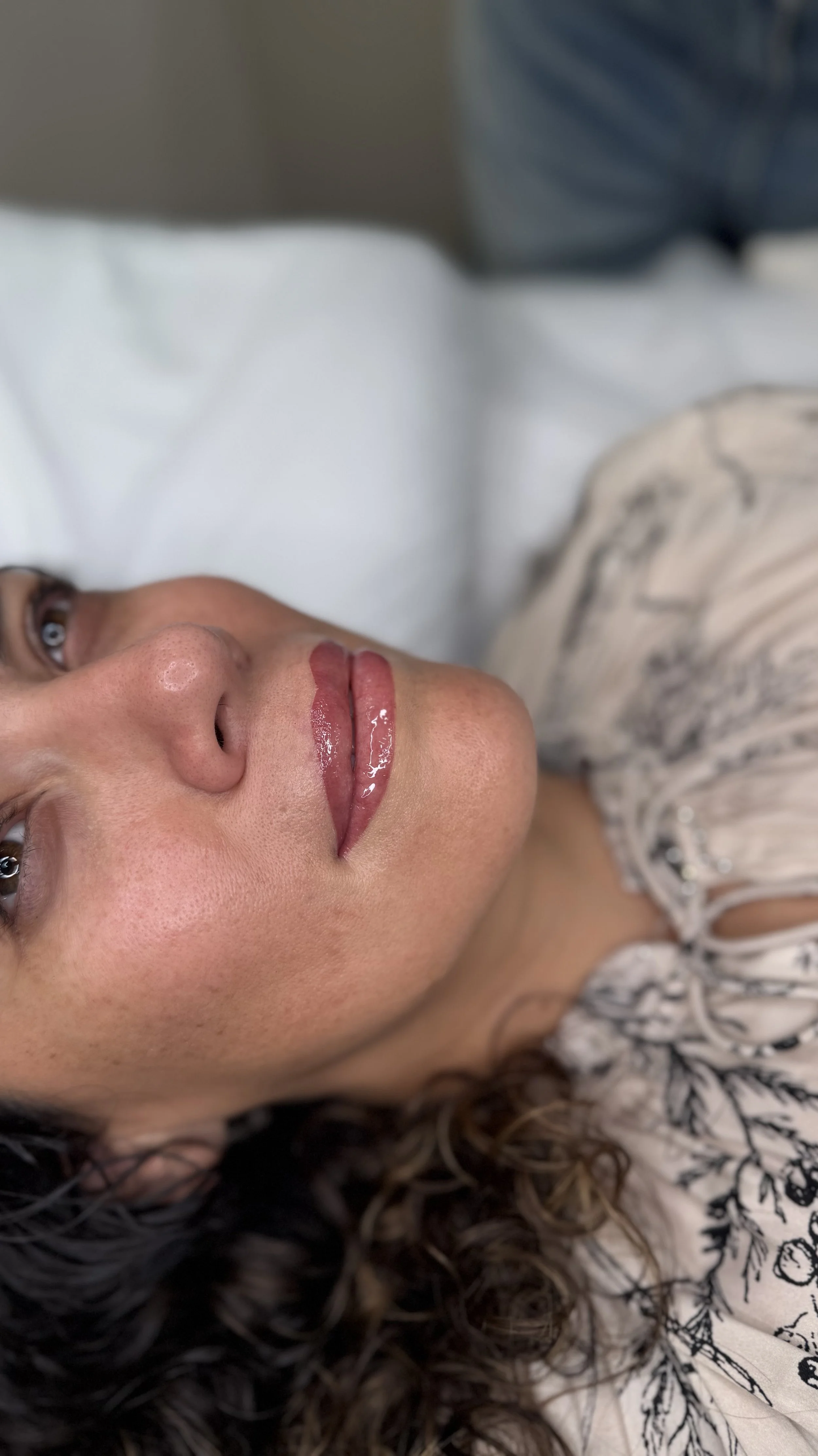 Close-up of a woman's face lying on a bed with curly hair, wearing a beige and black patterned top.