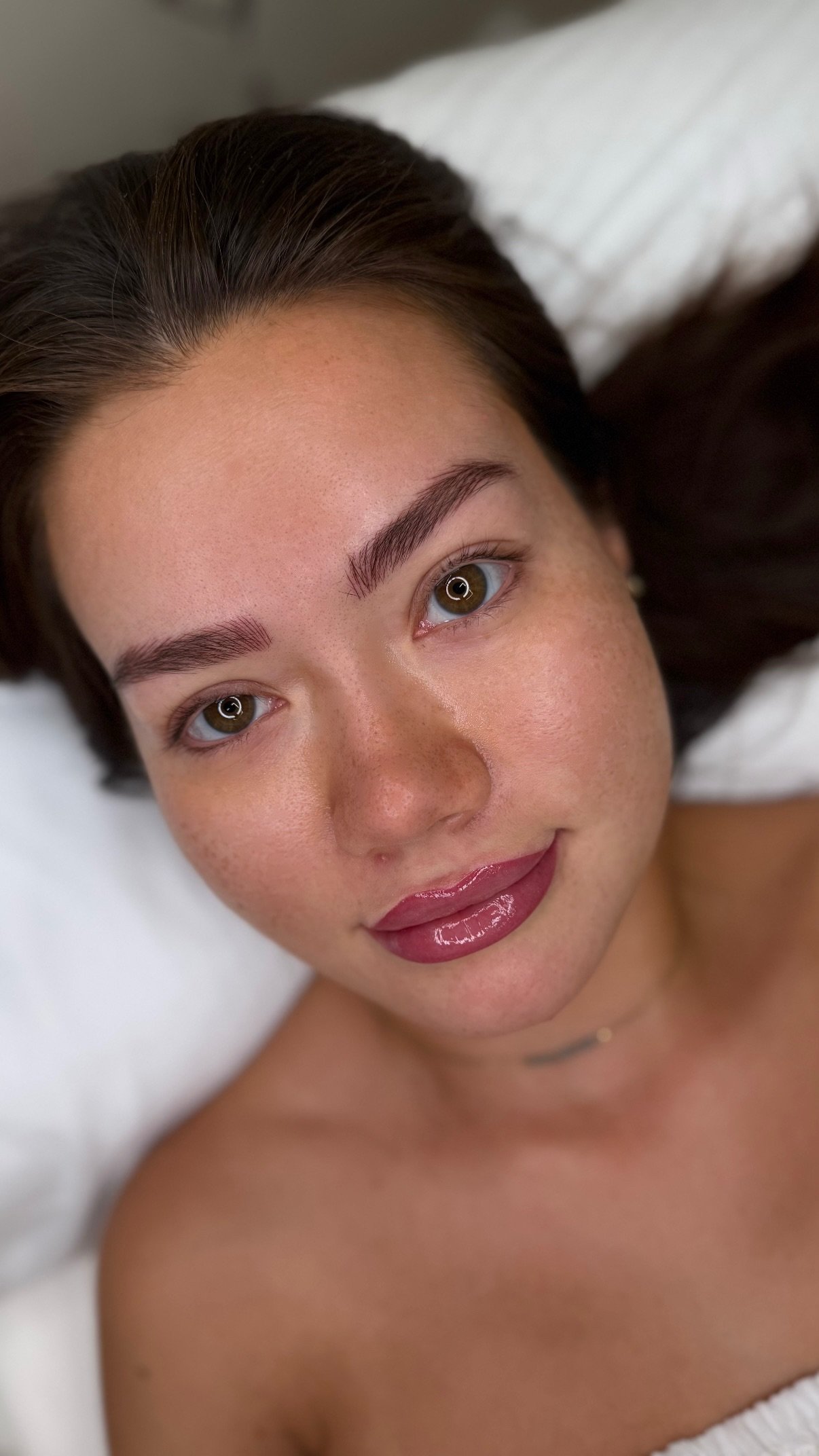 Close-up of a woman's face with brown hair, hazel eyes, smooth skin, and glossy lips, lying on a white pillow.